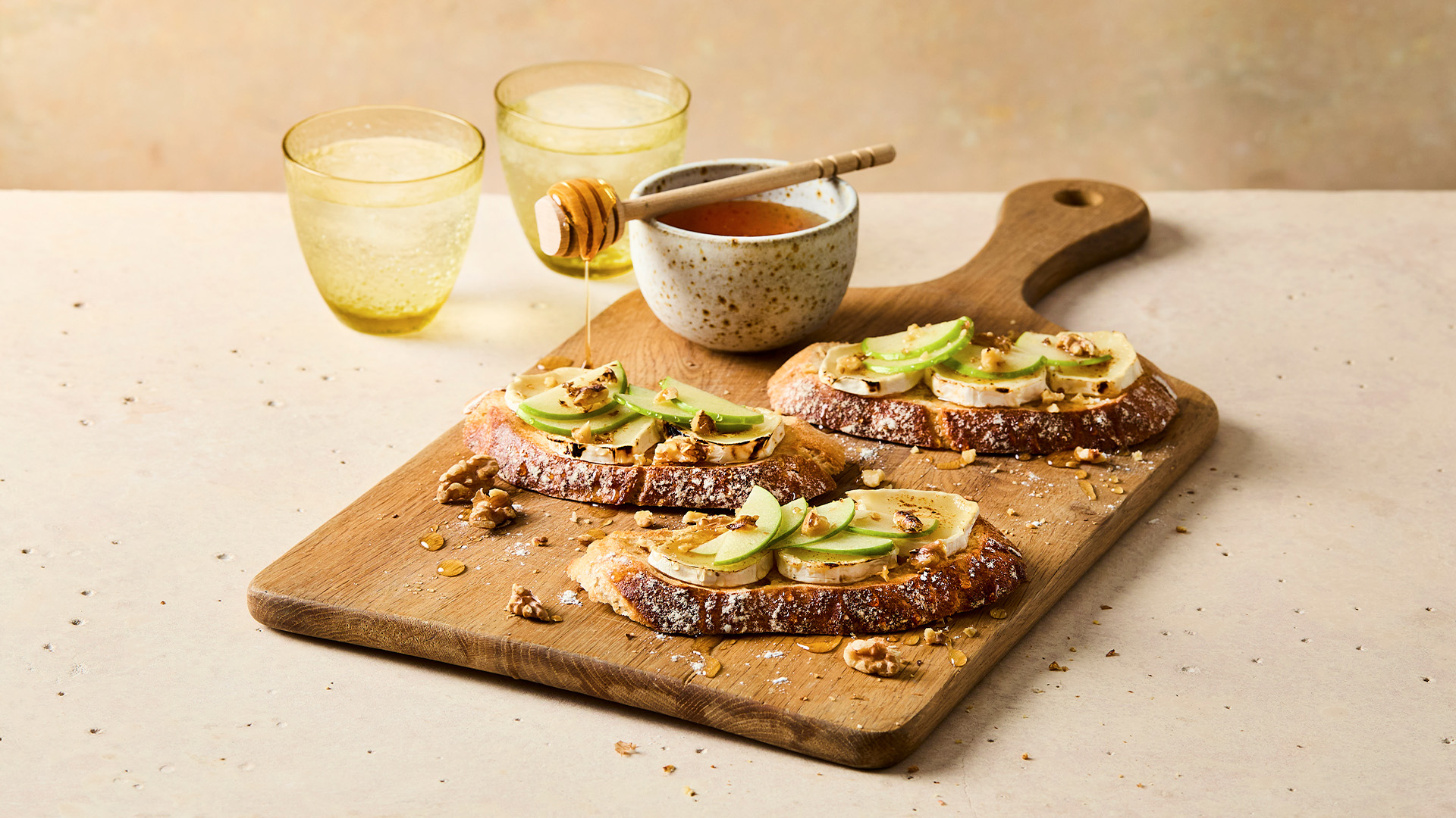 Three slices of seeded sourdough on a wooden chopping board, topped with goats cheese, apple and walnut pieces. there is a pot of honey dripping onto the chopping board behind the toasts. In the background there are two glasses of water.