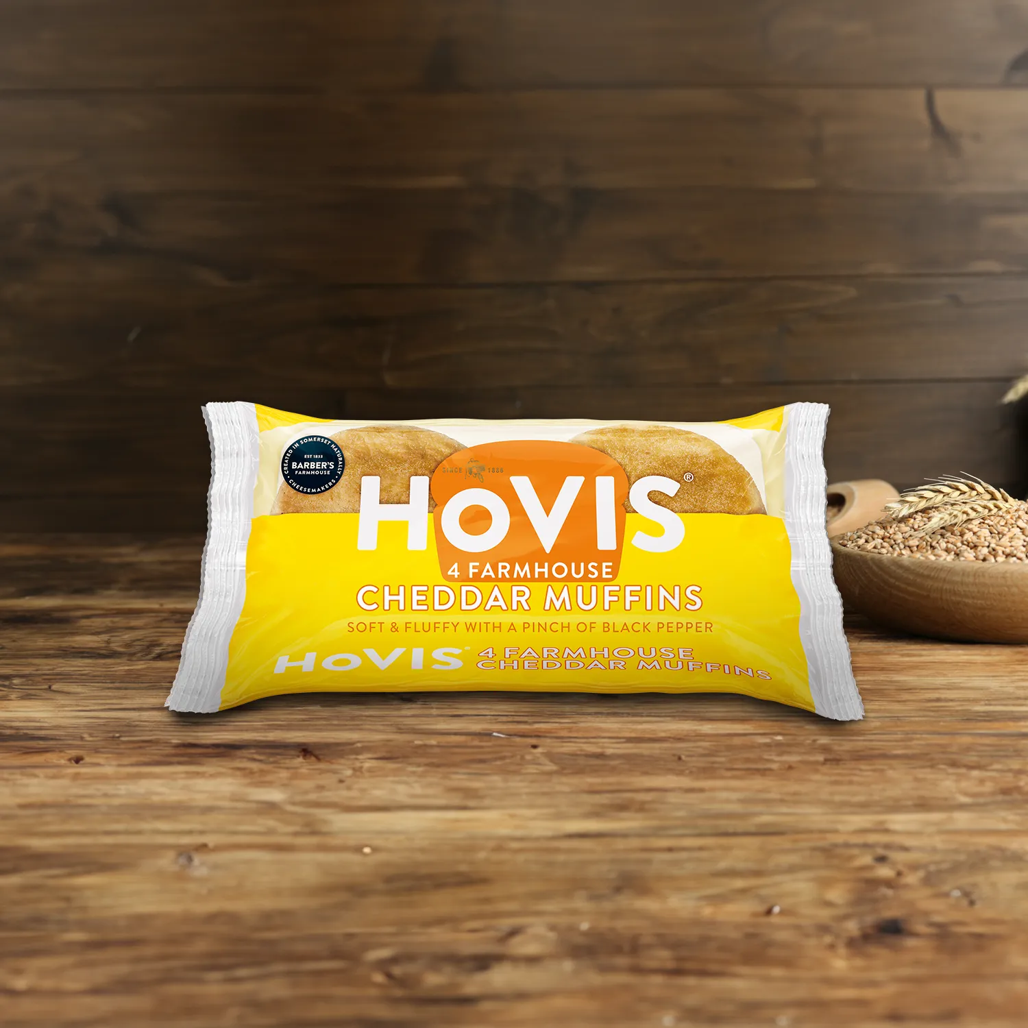 A pack of Hovis Farmhouse Cheddar Muffins on a wooden table, with a bowl of wheat grains, a wooden scoop, and dried wheat in the background.