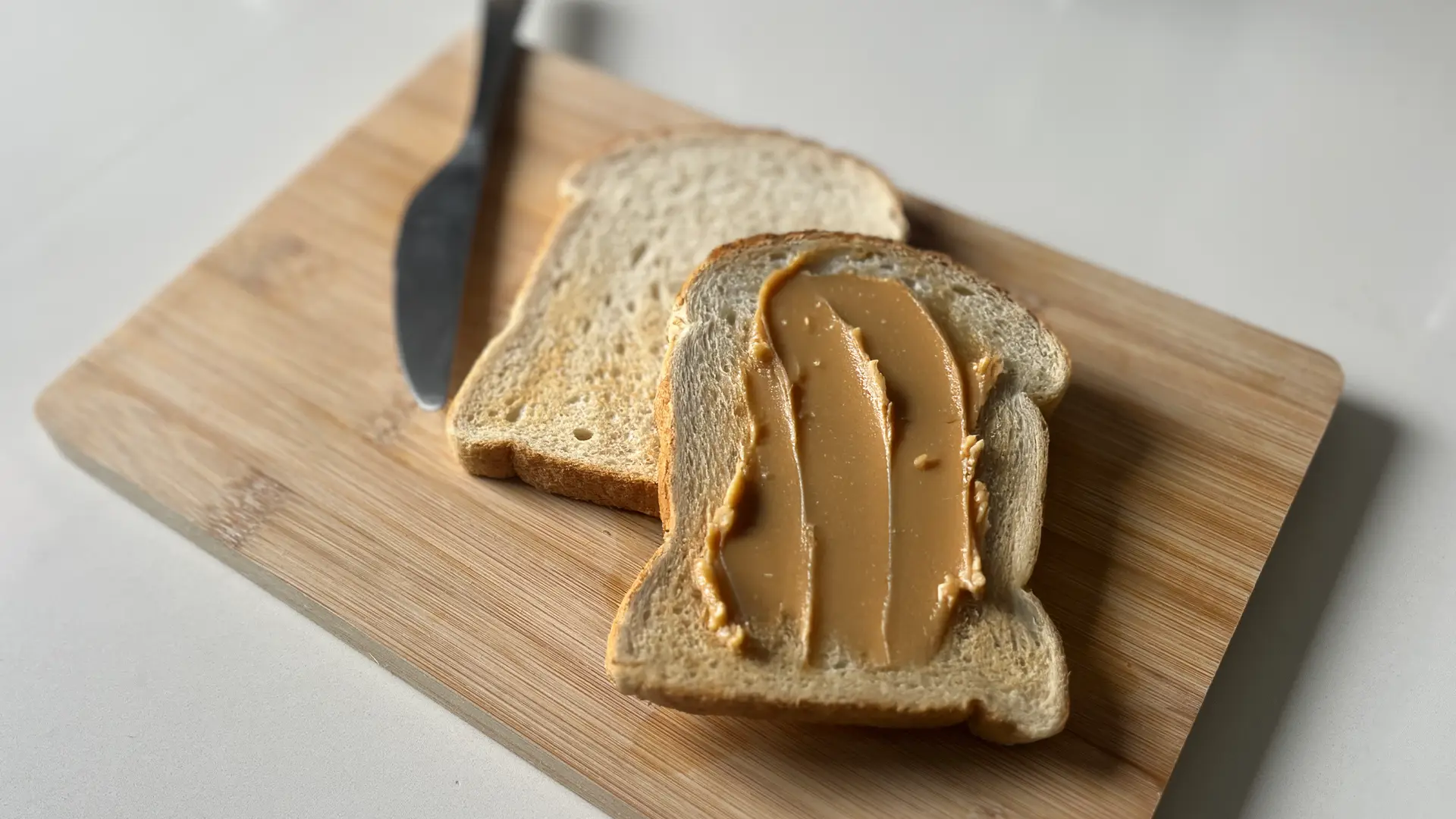 A top-down view shows two slices of lightly toasted white bread on a light wooden cutting board. The front slice is generously spread with smooth peanut butter, showing distinct knife marks. The back slice is plain. A silver butter knife rests on the board above the plain toast. The cutting board is positioned on a plain white surface.