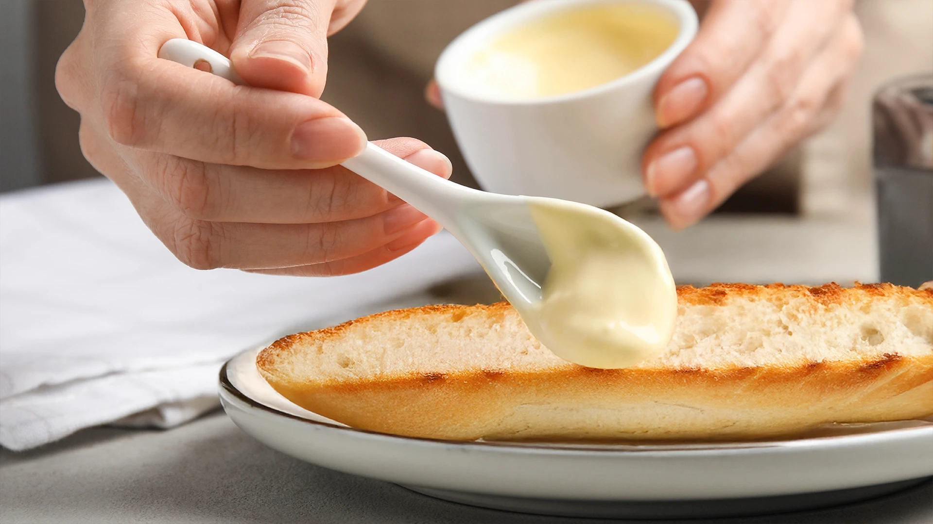 Someone spreading mayonnaise onto a half sub roll with a spoon