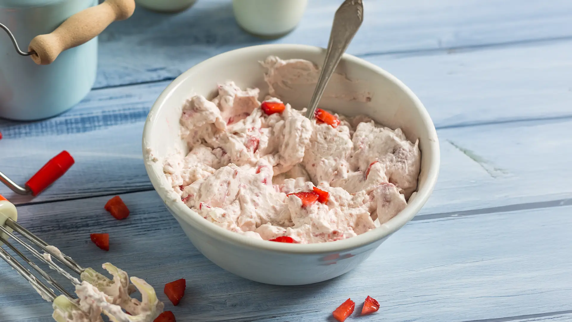 A white bowl filled with cream cheese and strawberry chunks sits on a blue wooden kitchen counter. Bits of strawberry are scattered around the bowl, and a whisk with cream cheese residue rests beside it.