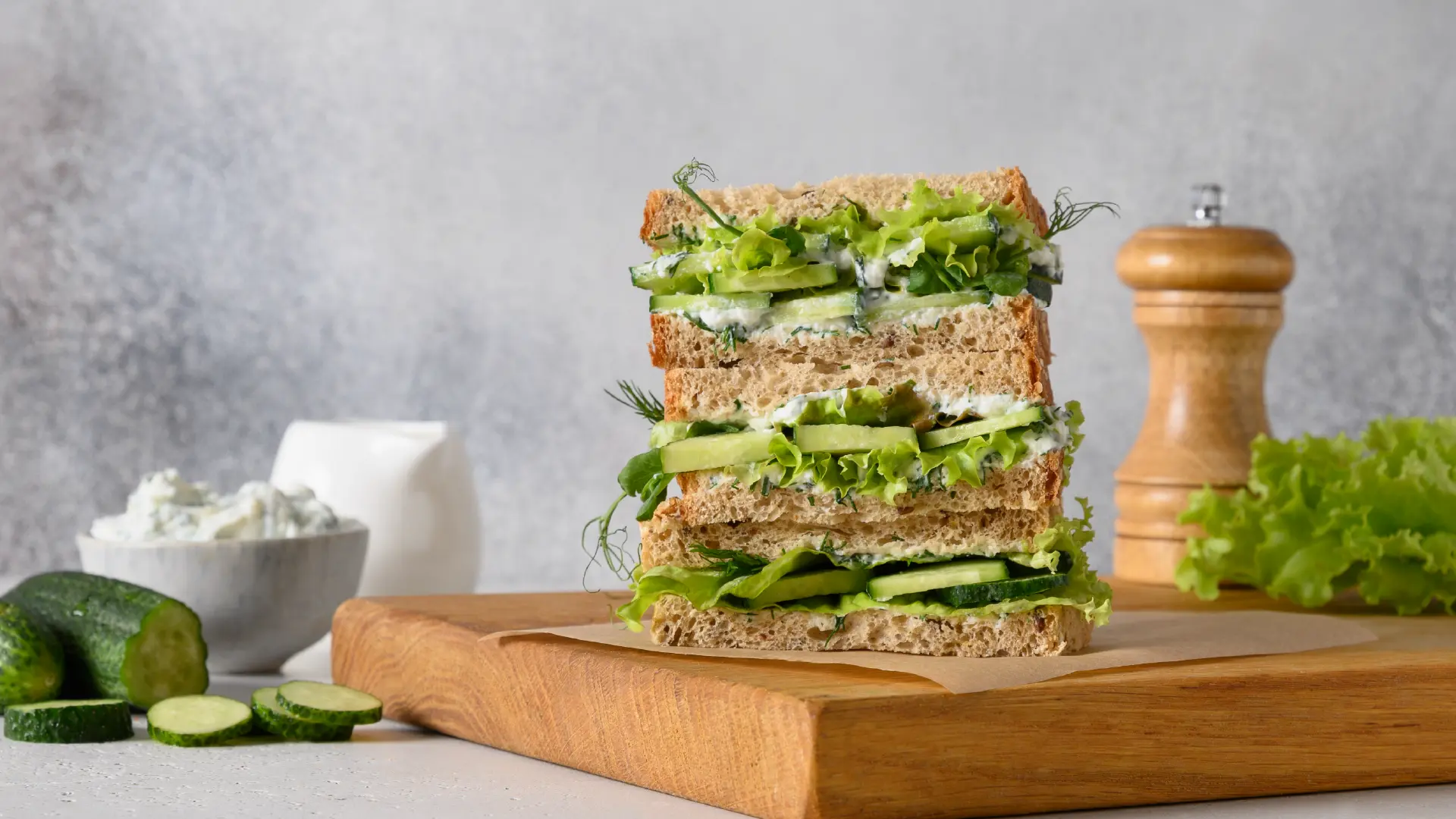 Three sandwiches made with Hovis Seeded Batch stacked on a wooden board, surrounded by a small bowl of cream cheese and sliced cucumber. Salad leaves, cucumber slices, cream cheese, and dill are spilling out from between the bread slices.