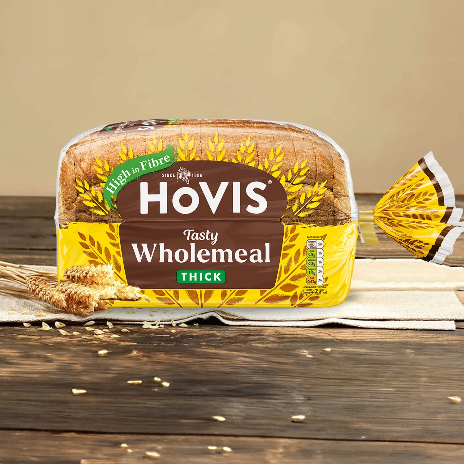 Hovis Tasty Wholemeal Thick Sliced bread on a light beige background atop a wooden table, surrounded by dried wheats.