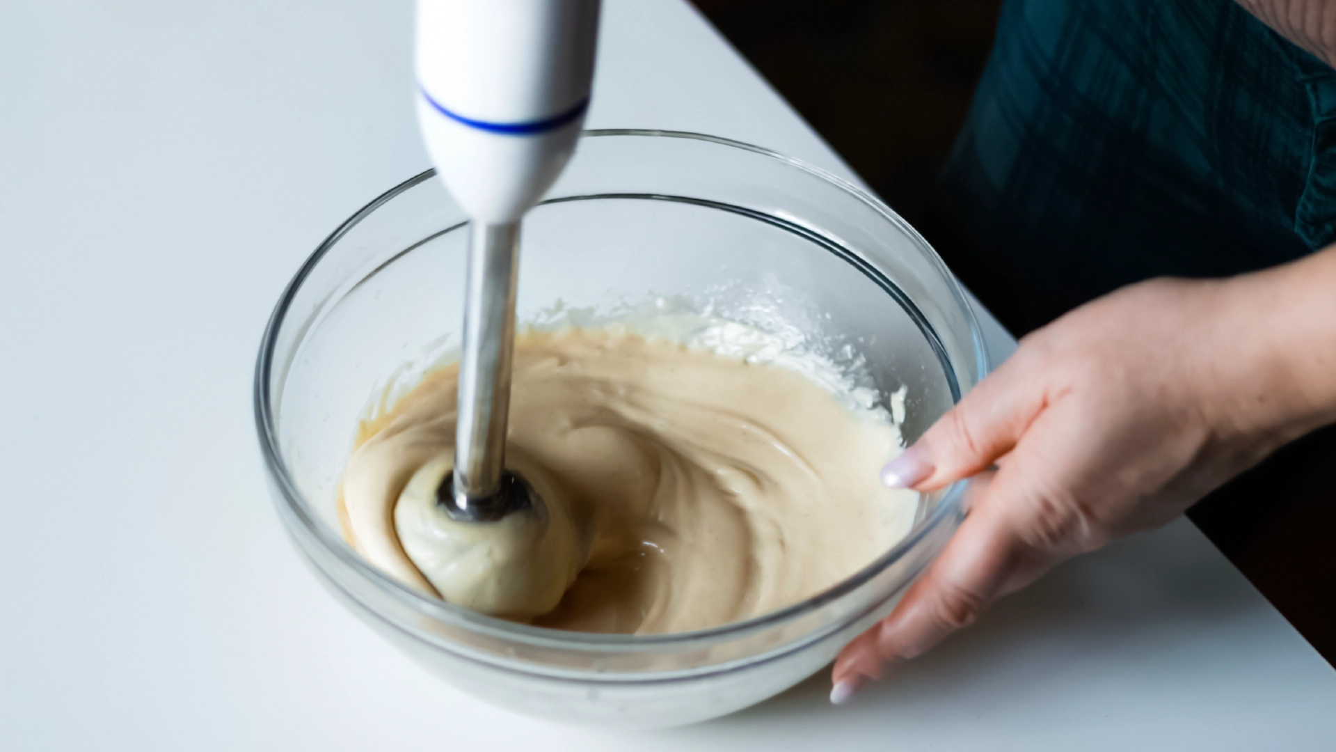 One hand holding a medium glass bowl while the other uses a hand blender to mix the mayonnaise on a white kitchen counter.