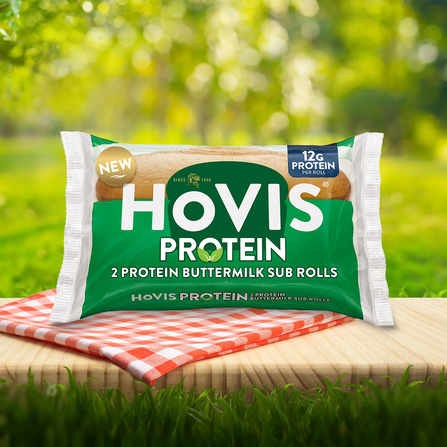 Hovis Protein Buttermilk Sub Rolls packaging on a picnic table with a green outdoor background. The pack shows two soft sub rolls with 12g of protein per roll and highlights “Deliciously soft and flavourful with a hint of buttermilk.” A red gingham cloth adds a picnic feel to the setting.