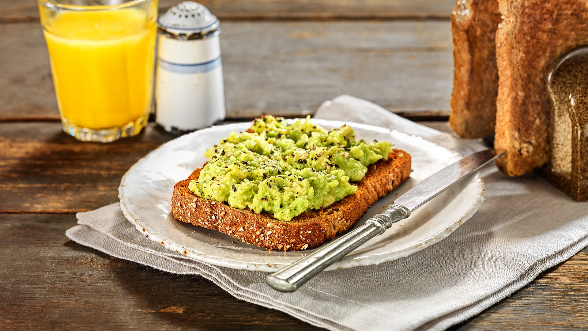 A slice of Hovis Granary Wholemeal bread topped with smashed avocado and a sprinkle of ground black pepper, on a plate with a butter knife on the side.