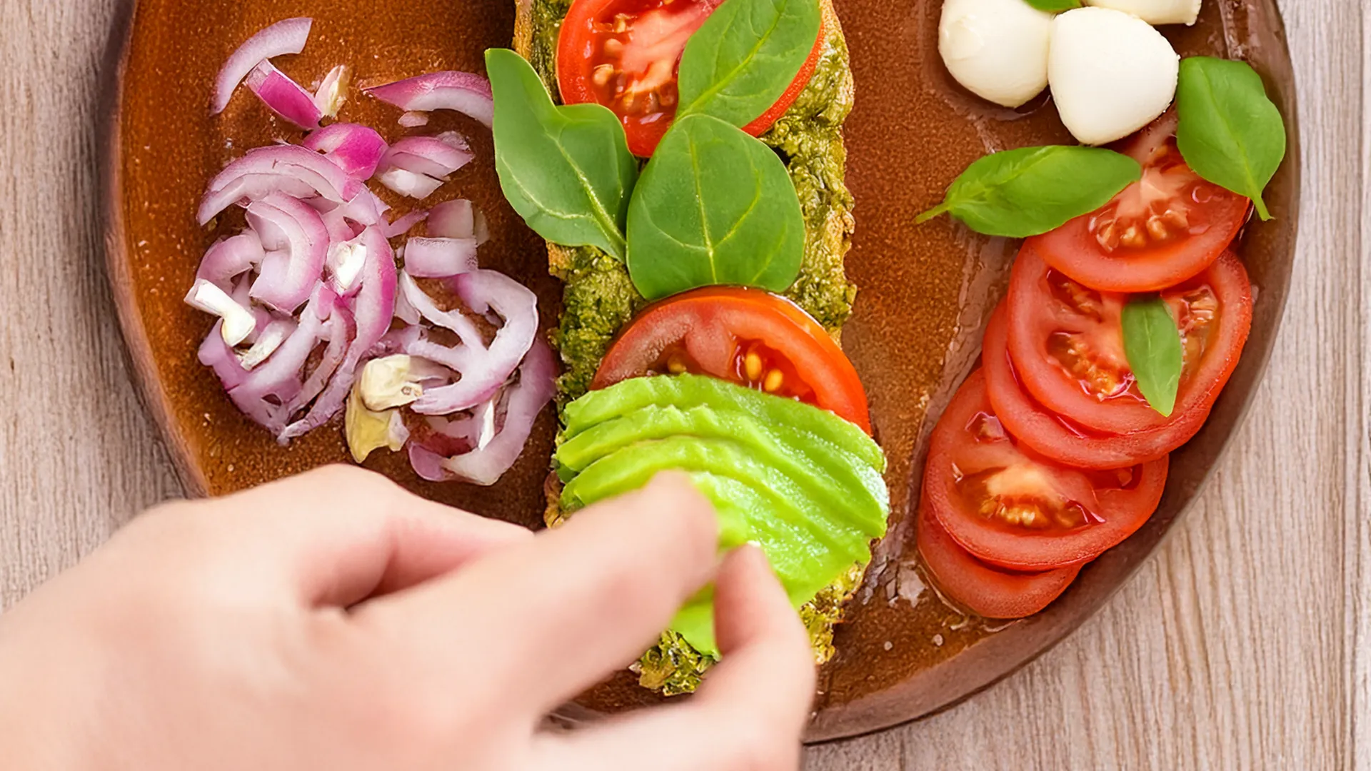 the bottom half of a sub roll sits on a red plate with pesto, tomato and spinach already on top. Next to it, more tomato slices, mozzarella balls and chopped red onion wait to be placed as someone is starting to place avocado on the roll