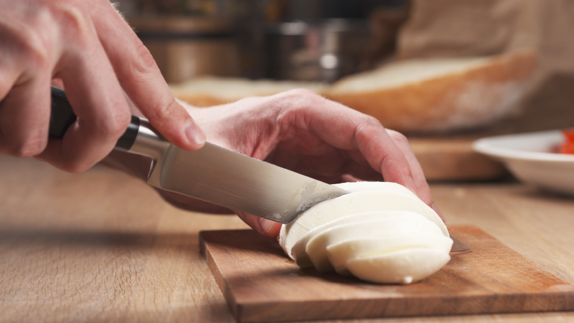 A hand holds a mozzarella ball while the other uses a sharp knife to slice it into thinner pieces on a wooden chopping board.
