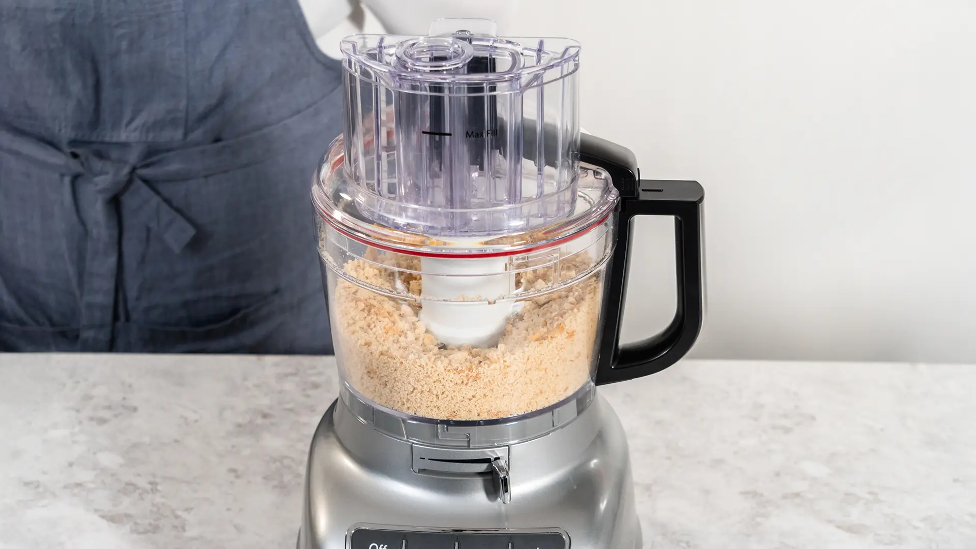 A food processor is on top of a white marble counter. Inside are small pieces of crumbled bread. A person wearing a dark blue apron can be seen in the background. 