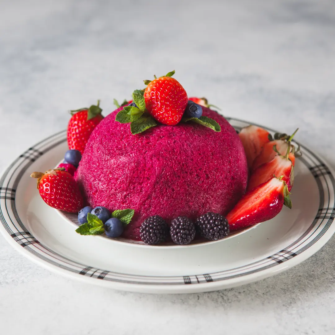 A small, bright pink dome-shaped bread pudding made with Hovis Farmhouse Soft White on a plate, topped with a strawberry and mint leaves. The base is surrounded by halved strawberries, blackberries, blueberries, and more mint leaves.