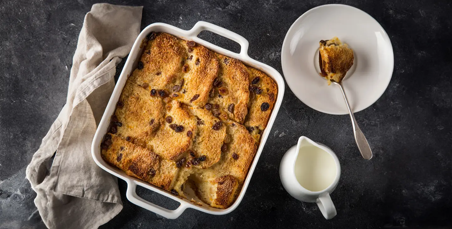 A Bread and Butter pudding sits in an oven proof dish on a kitchen table surrounded by cream and a plate. 