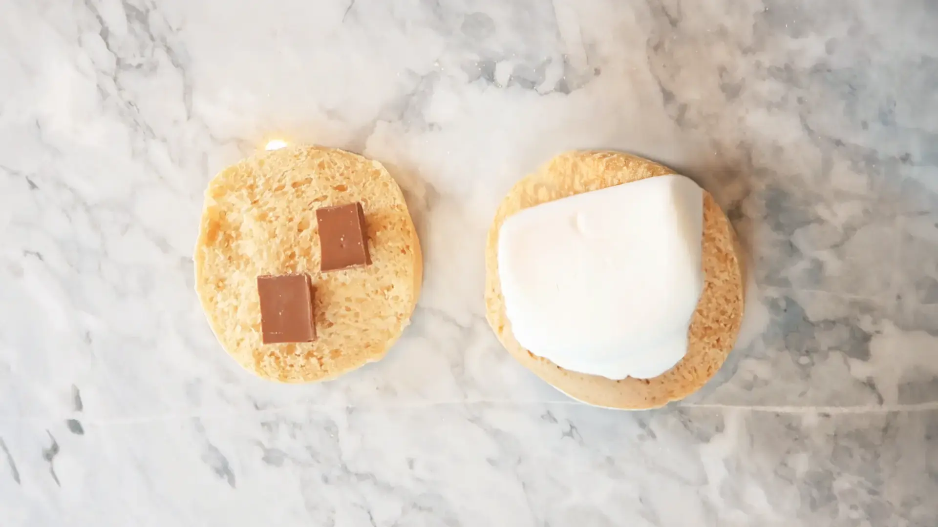 A top-down view of a Hovis English Muffin sliced in half on top of a marbled counter. On top of the slice to the left there are two small chocolate squares. On top of the slice to the right, marshmallows.