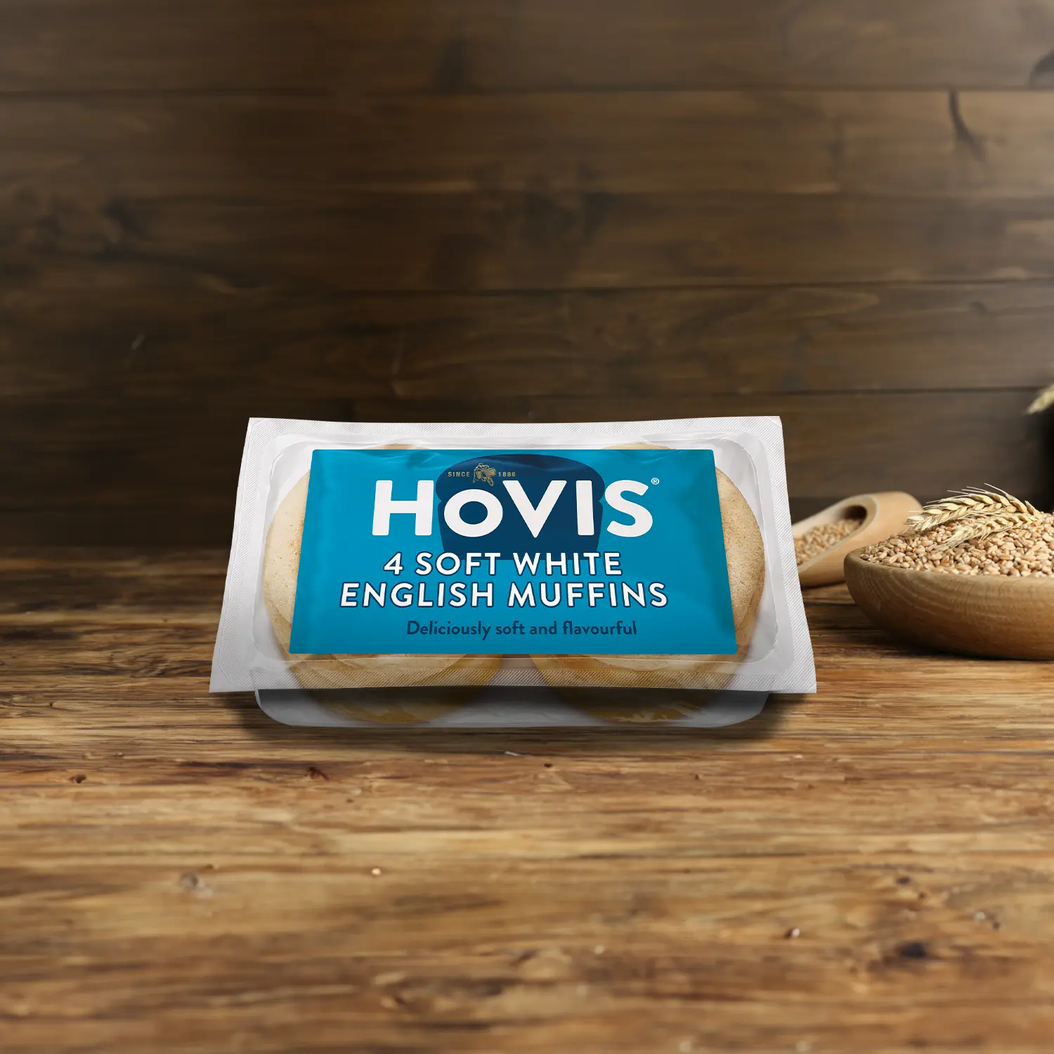 A pack four Hovis Soft White Muffins on a wooden table, with a bowl of wheat grains, a wooden scoop, and dried wheat in the background.