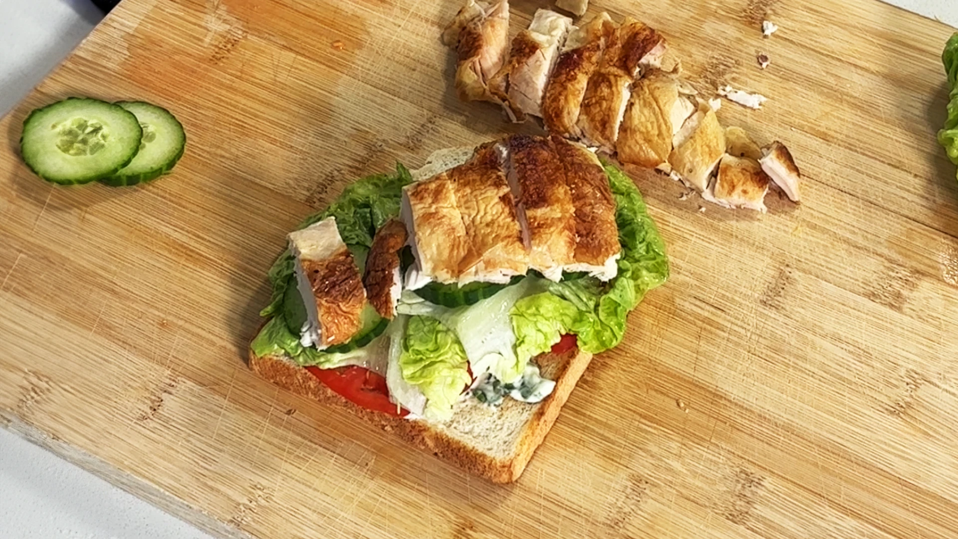 One slice of Hovis Soft White Medium Sliced bread topped with sliced tomatoes, salad leaves, and sliced chicken on a wooden chopping board. Some sliced chicken and cucumber on the side.