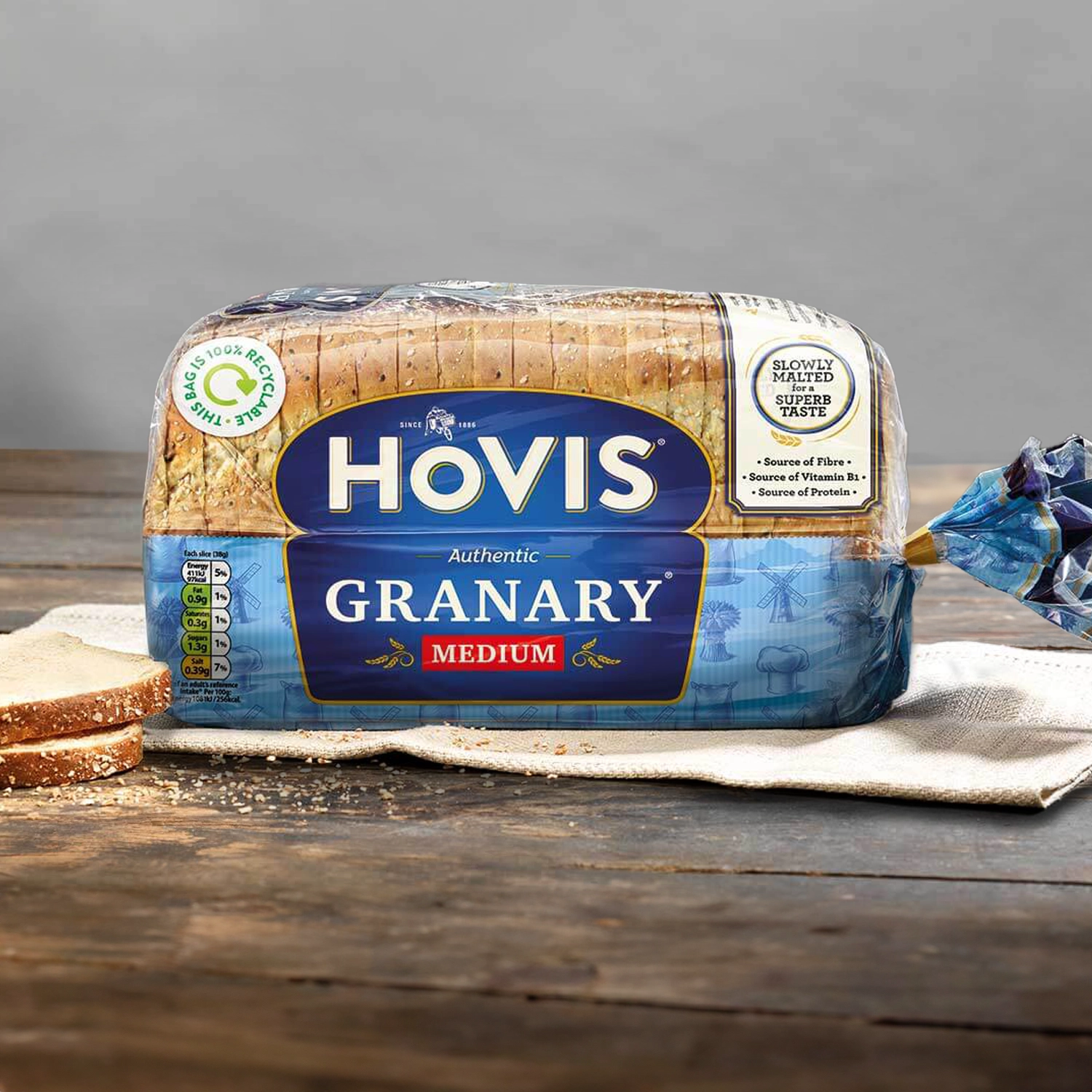 Hovis Granary Medium Sliced loaf on beige napkins against a light grey background, resting on a rustic wooden table with natural light.