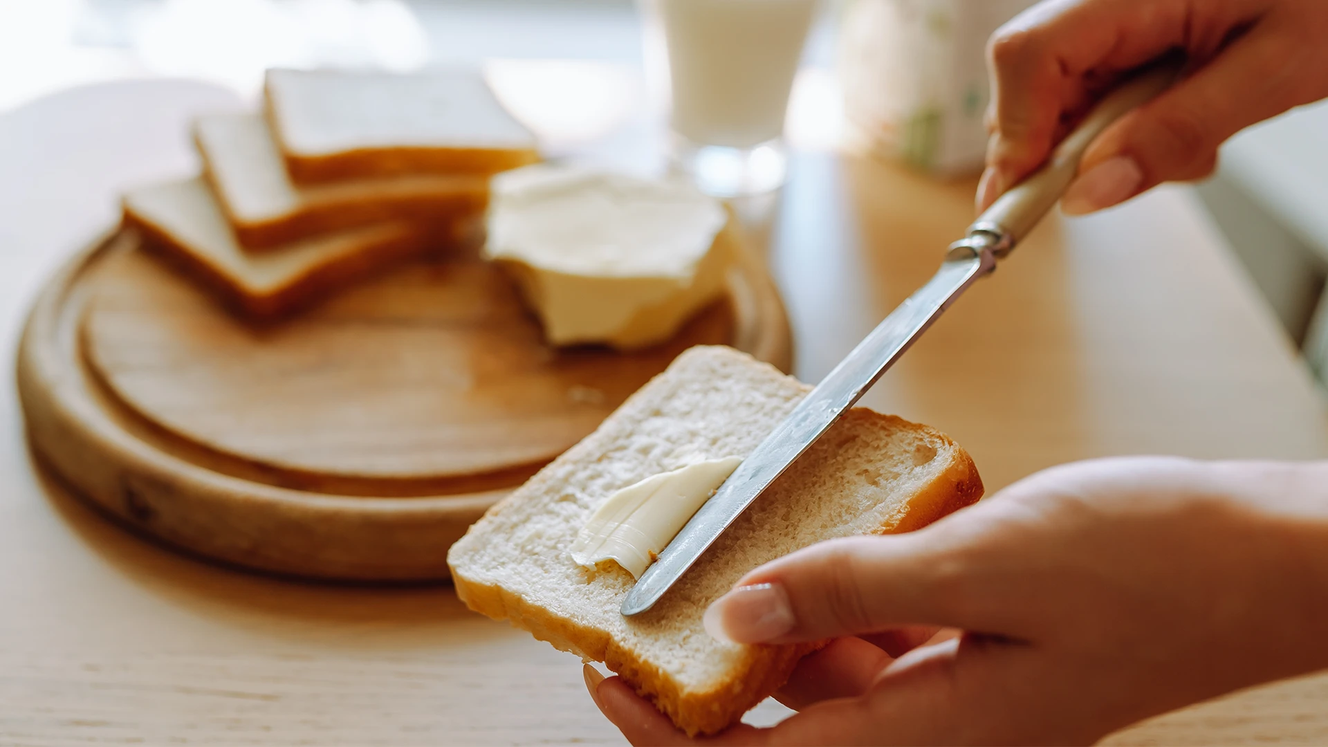 A slice of Hovis Soft White Medium bread being buttered with a knife, with three more stacked slices on a wooden chopping board in the background.