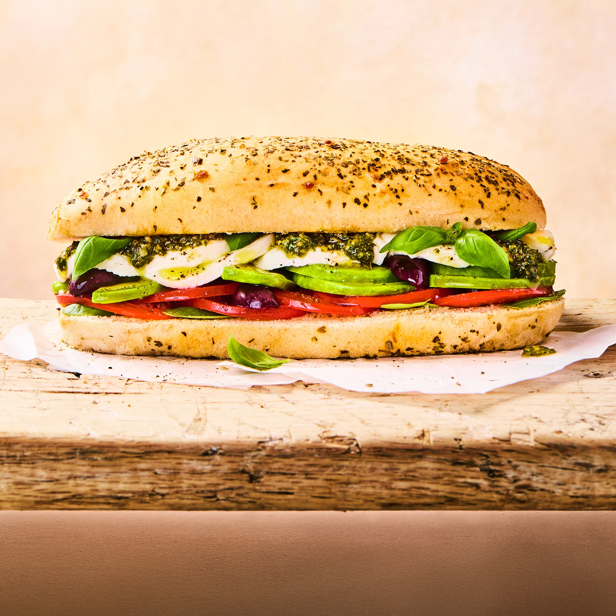 A cheese and herb sub roll sitting on parchment paper on a wooden surface with the tomato, pesto, spinach, avocado and mozzarella toppings peeking out