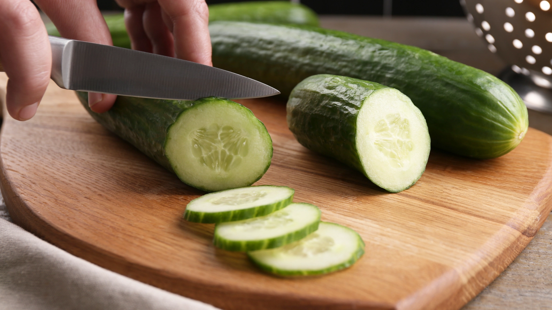 Hands using a small, sharp knife to thinly slice a fresh cucumber on a wooden chopping board, with additional cucumbers in the background.