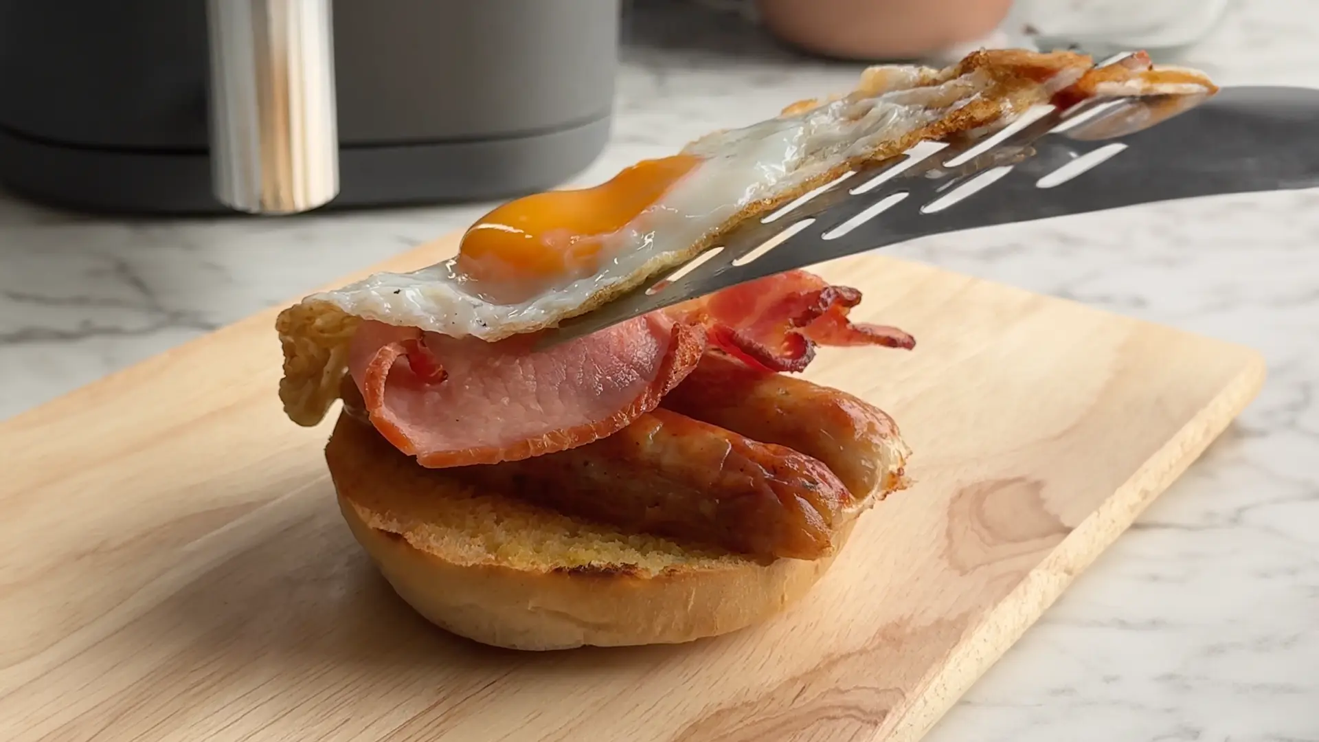 Someone uses a metal spatula to place an egg with a runny yolk on top of crispy bacon, sausage and toasted Hovis Burger Bun.