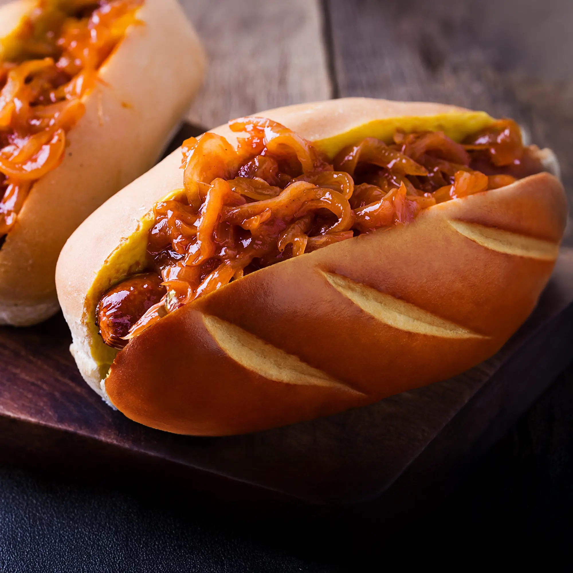 A delicious hot dog in a lightly toasted bun, generously topped with an abundance of glistening caramelized onions. The bun has decorative diagonal slashes on its golden-brown crust. A hint of another hot dog is visible in the blurred background on the left. The hot dog and bun are resting on a rustic wooden board, with a dark, blurred background.