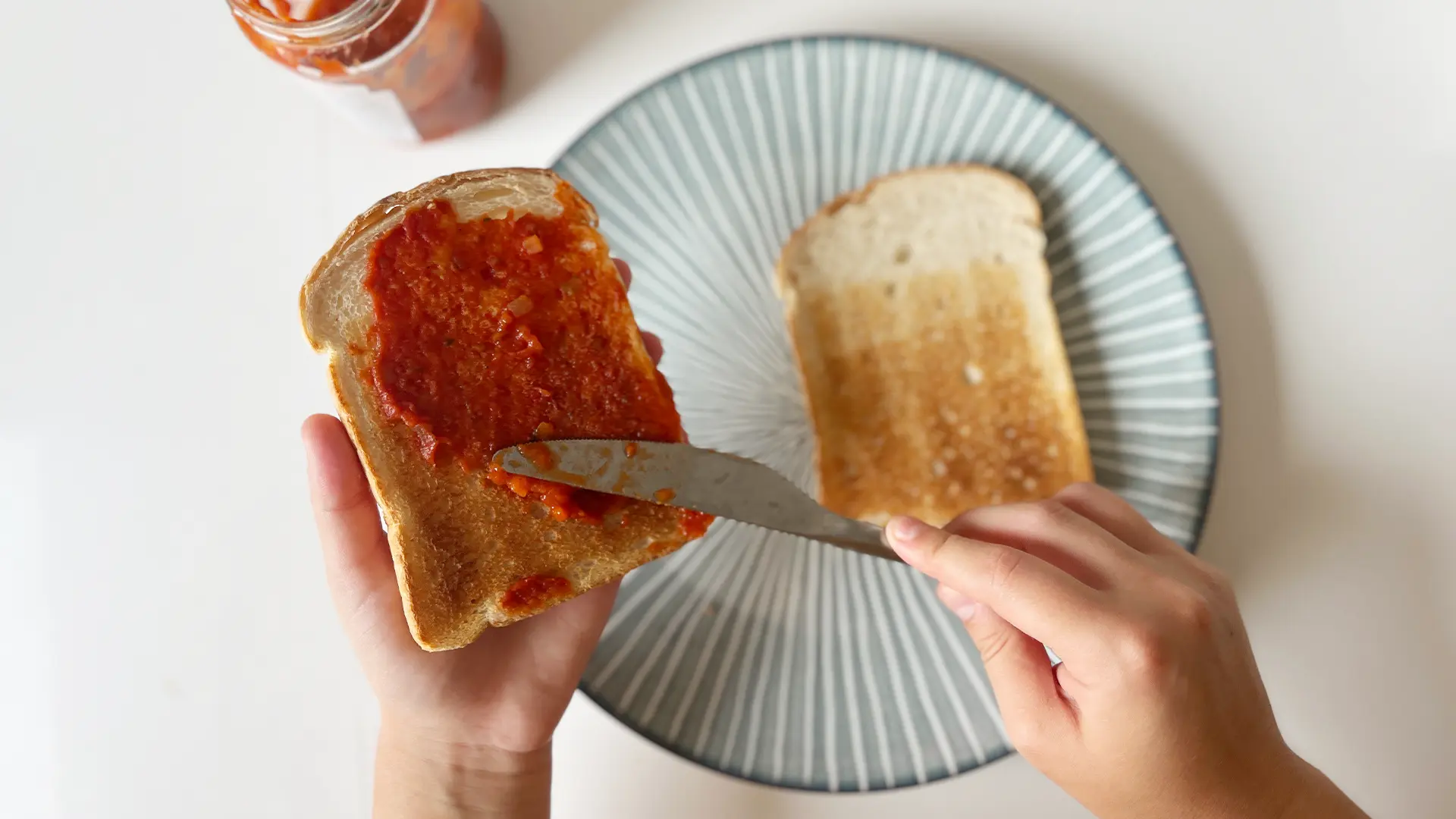A close-up image of tomato paste being spread on a slice of Toasted Hovis® White ‘n Fibre Bread with a jar of sauce in the background