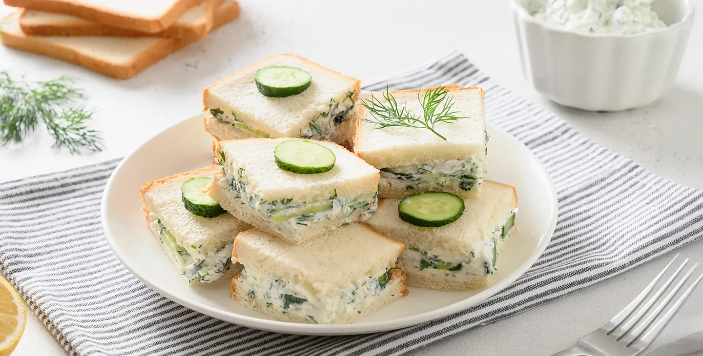 A bright, overhead shot of a plate piled high with six perfectly cut cucumber sandwiches, likely for afternoon tea. The sandwiches are made with white bread, have their crusts removed, and are filled with a creamy green spread, visible cucumber slices, and fresh dill. Several are garnished with a thin slice of cucumber on top, while one features a small sprig of dill. The plate rests on a grey and white striped tea towel. In the background, stacks of plain white bread slices are visible, along with a lemon half, a sprig of fresh dill, a bowl of the creamy spread, and a fork and knife set.