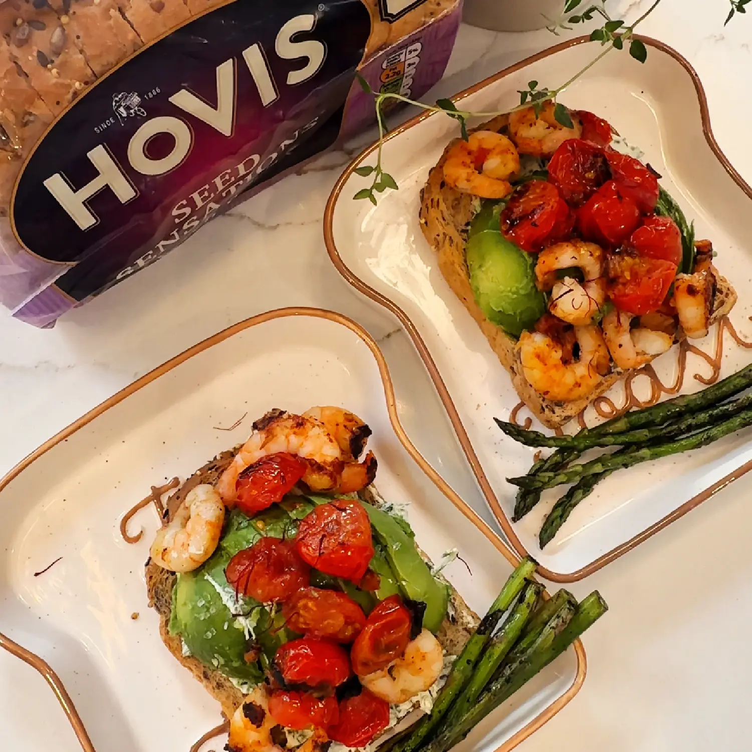 Two plates of seeded toast topped with avocado, grilled shrimp, and tomatoes, served with a side of charred asparagus.