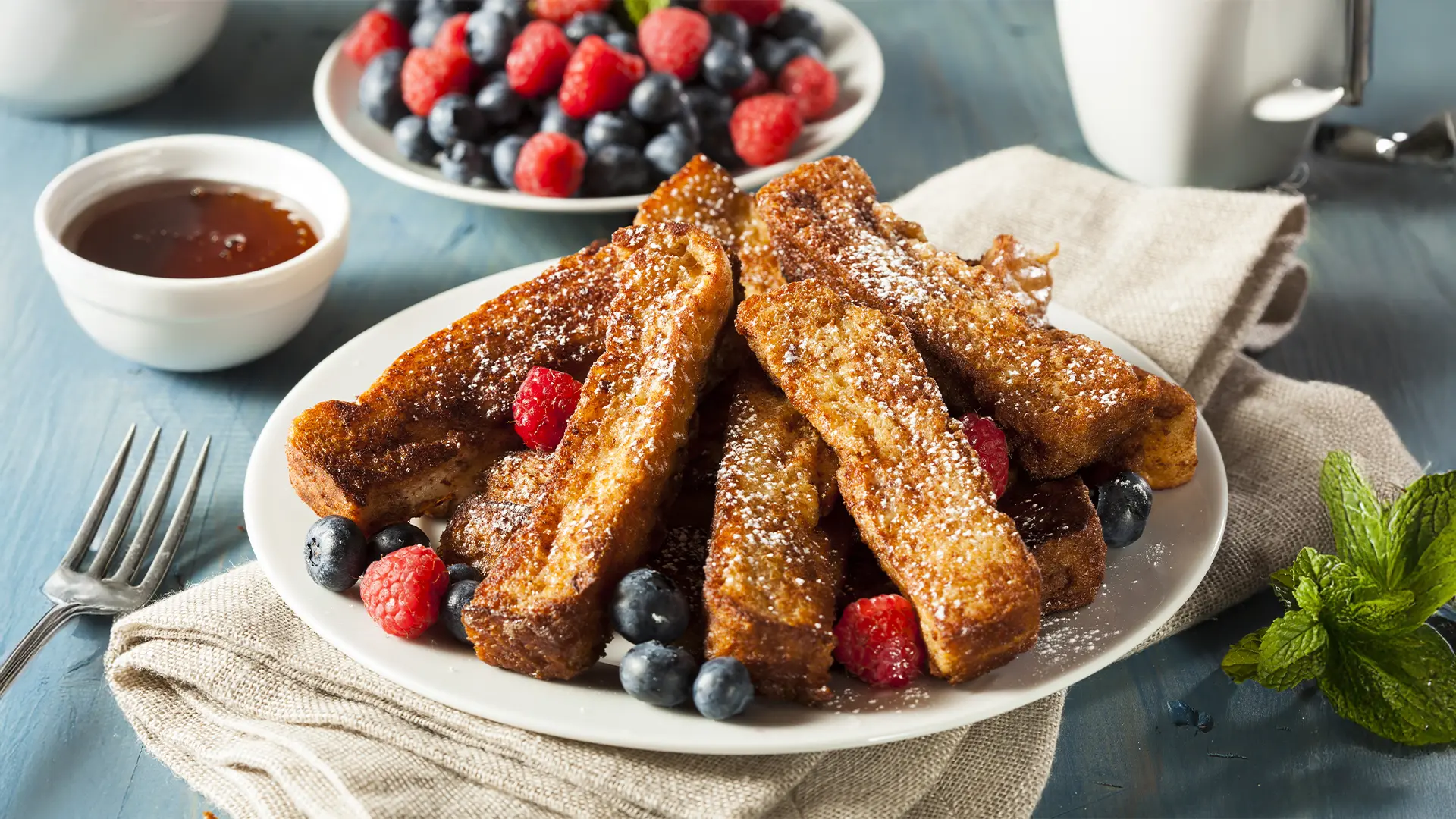  Golden brown bread sticks topped with sugar, surrounded by berries on a white plate. In the background some extra berries and a small bowl with chocolate dip. 