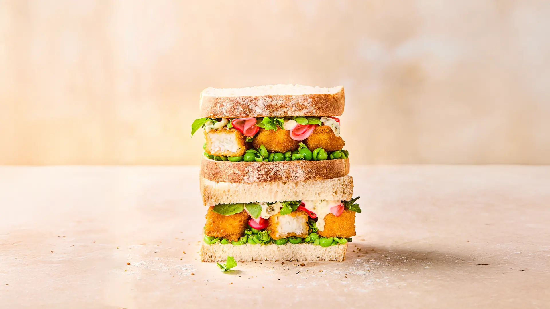 Two Hovis White n Fibre sandwiches, stacked, filled with golden fish fingers, tartar sauce, pickled radish, salad leaves and peas, resting on top of a table in a light beige background.