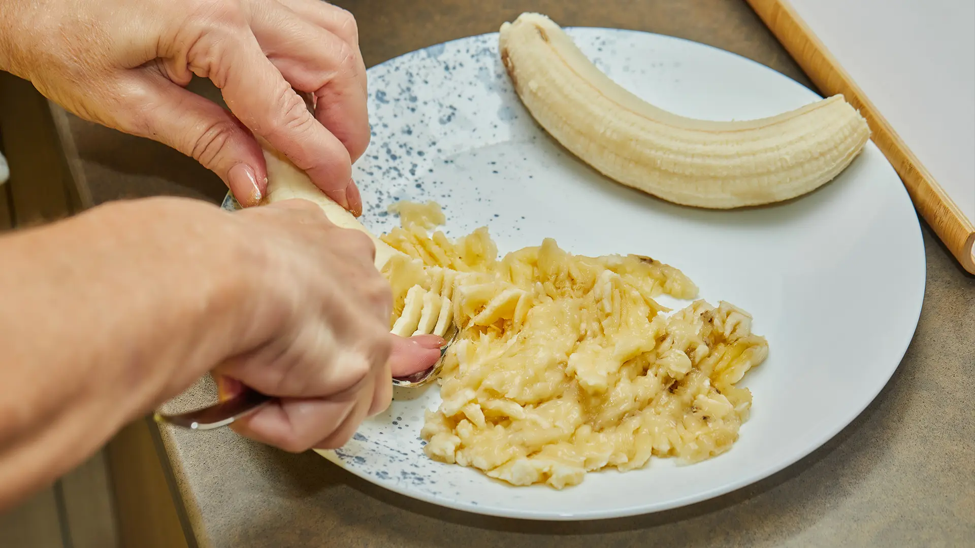 Close-up of hands mashing a banana with a fork on a white plate, with half a peeled banana beside the mashed portion.