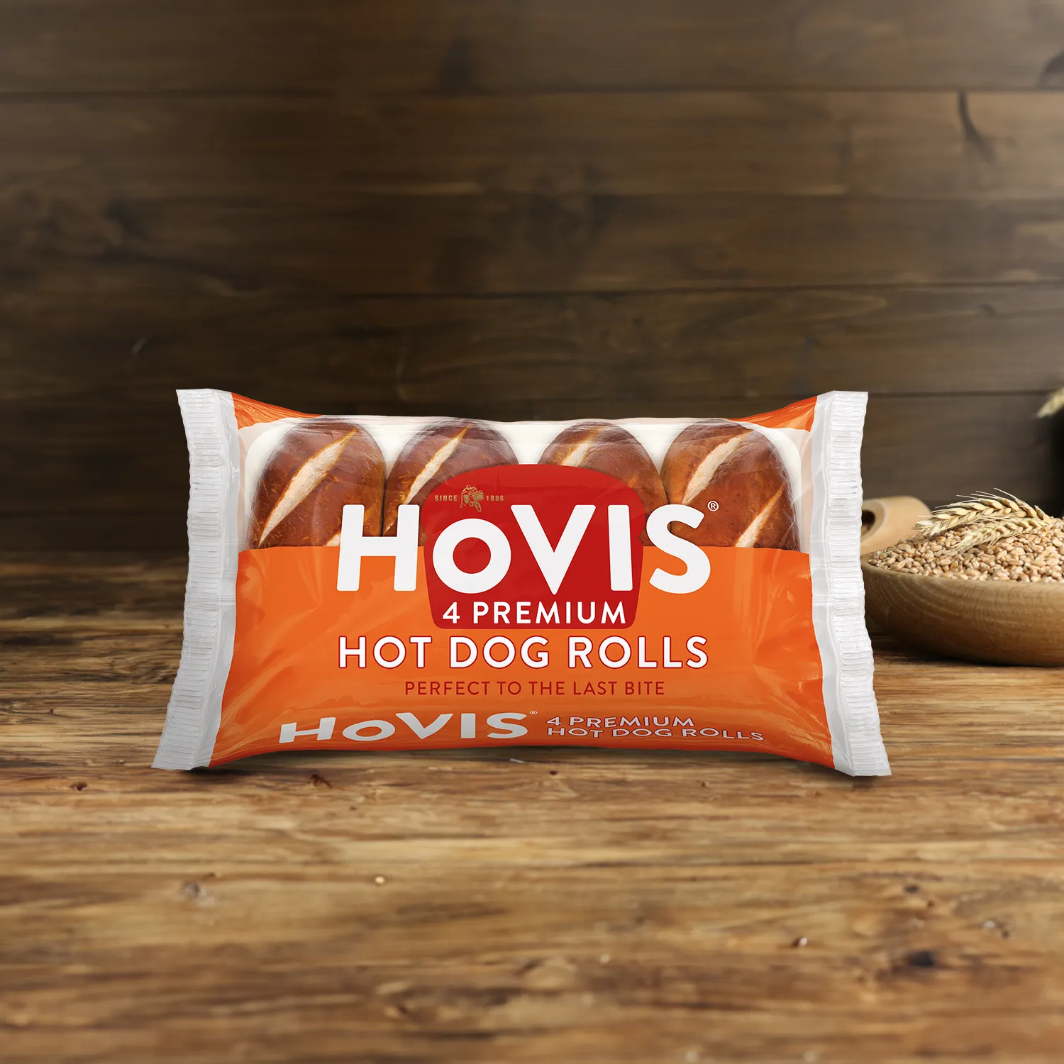Four Hovis Hot Dog Rolls on a wooden table, with a bowl of wheat grains, a wooden scoop, and dried wheat in the background.