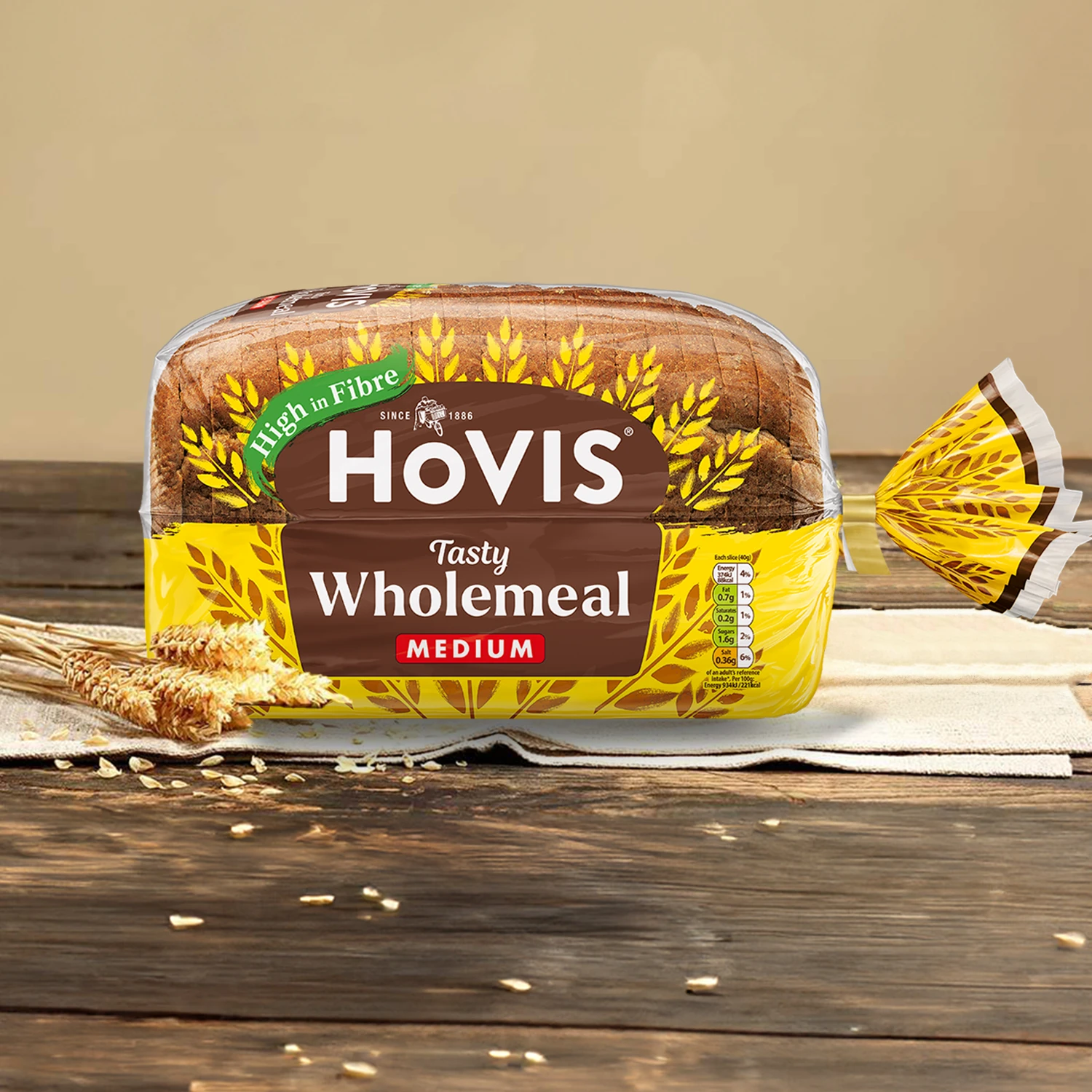 Hovis Tasty Wholemeal Medium Sliced bread on napkins against a light beige background atop a wooden table, surrounded by dried wheat.