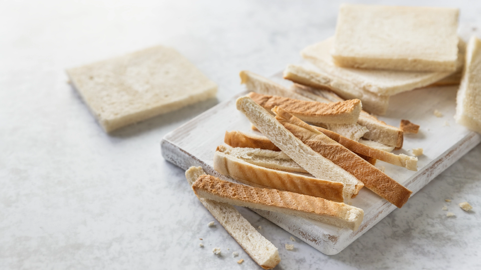 Slices of Hovis Farmhouse Batch bread with the crusts removed, placed on a cutting board with the crusts set aside, in a marbled background. 