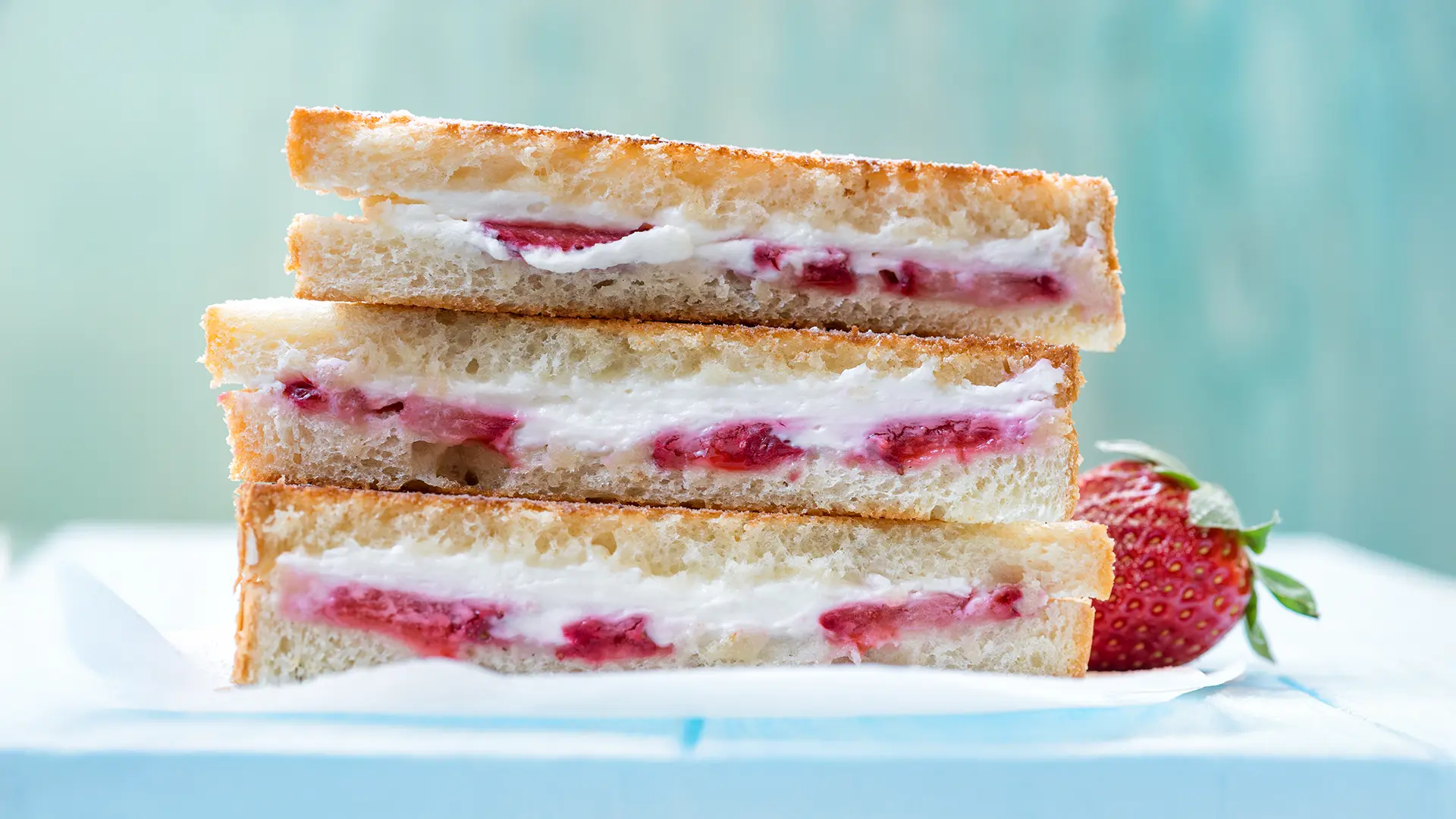 A delicious looking  strawberry-stuffed French toast sandwich, cut open to reveal its creamy strawberry filling, sits on a white surface with a whole strawberry beside it.