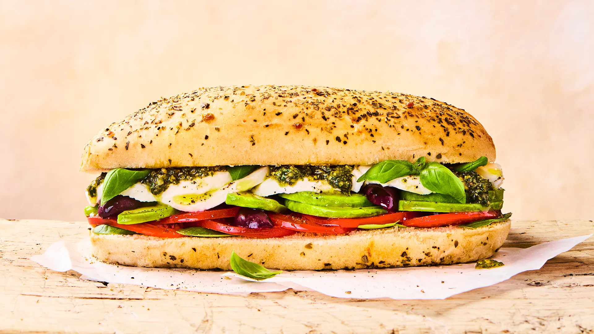 a complete Mozzarella, pesto, tomato and avocado sub sandwich sits on top of parchment paper on a wooden surface. the ingredients are clearly visible between the sub roll halves.