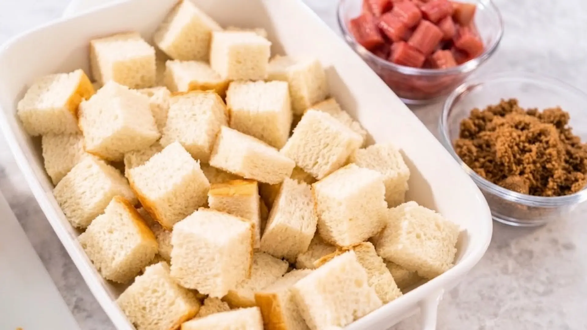 Diced bread cubes fill a white baking dish, with small bowls of chopped rhubarb and brown sugar sitting nearby.