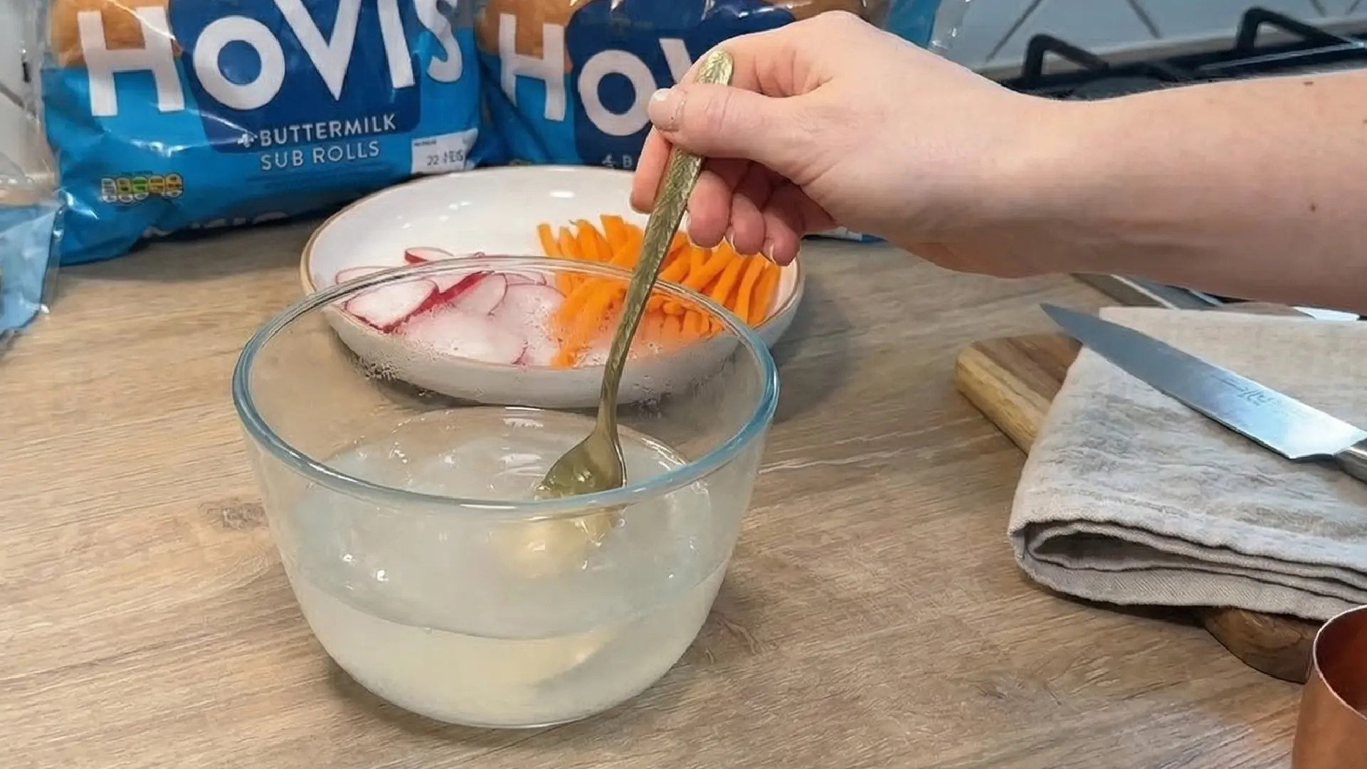 A hand stirs a clear bowl filled with a mixture of water, sugar and white vinegar. Hovis buttermilk sub rolls and a plate of raddish and carrots are in the background. 