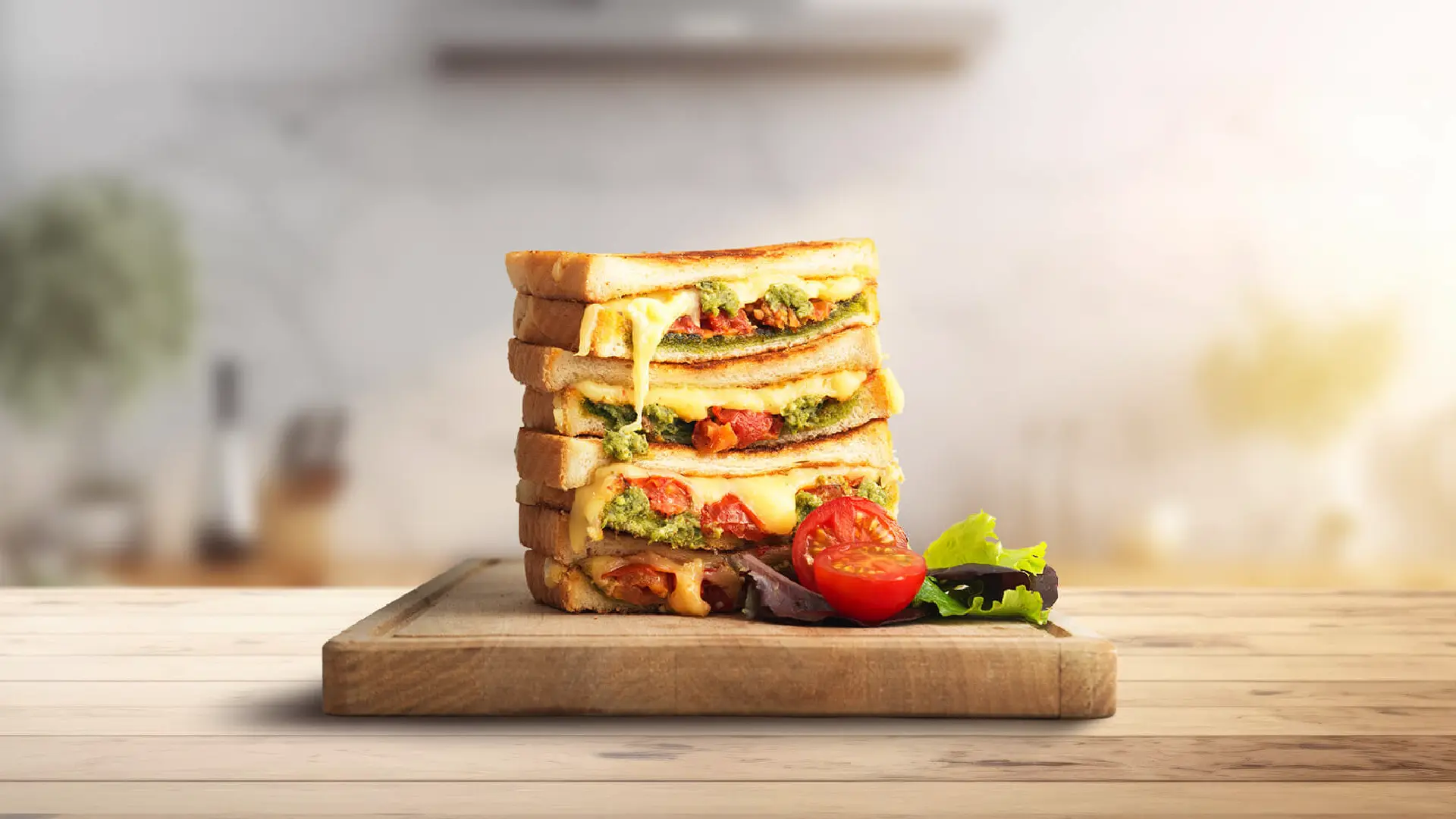 Four halves of pesto and cheese toasties made with Hovis Soft White Medium Sliced stacked on a wooden chopping board, with molten cheese and pesto slightly visible between the slices. A small halved tomato and some salad leaves are placed to the right.