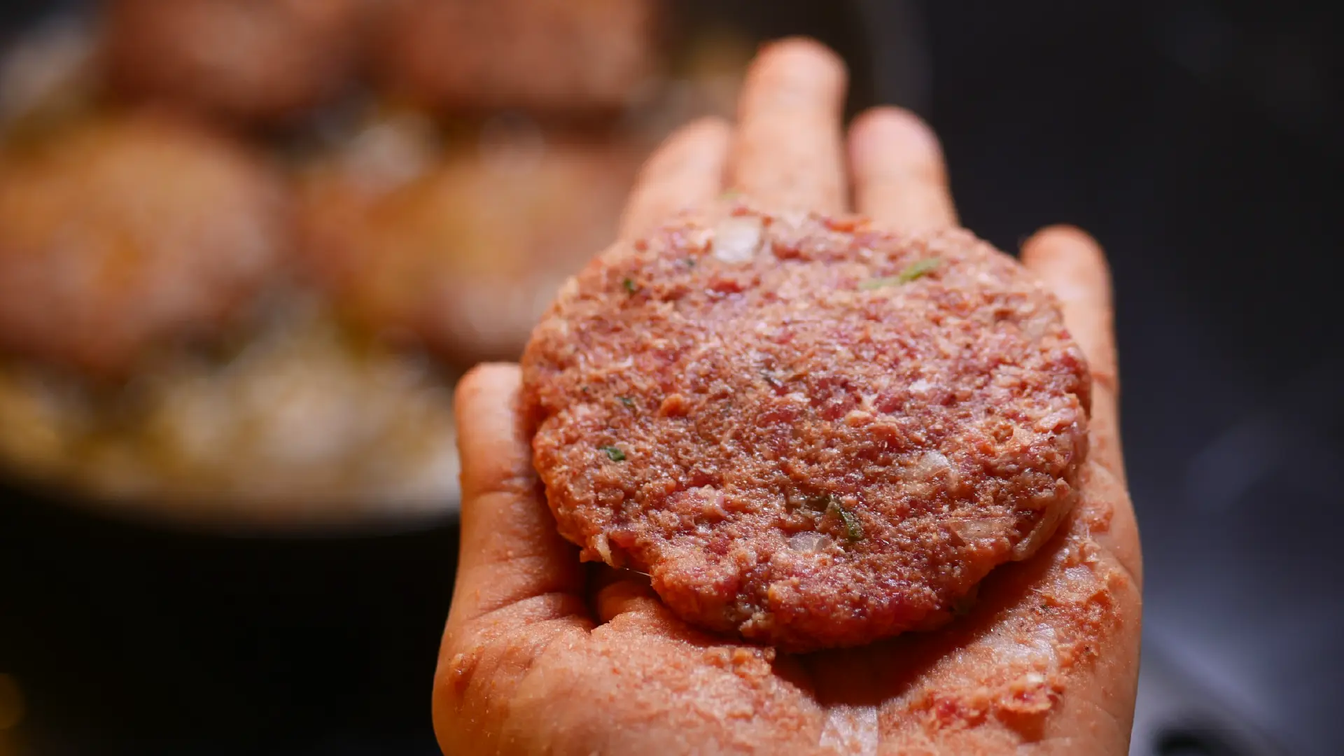 Someone holds a meat patty, with seasoning, in their hands. In the background, some extra patties can be seen in a frying pan.