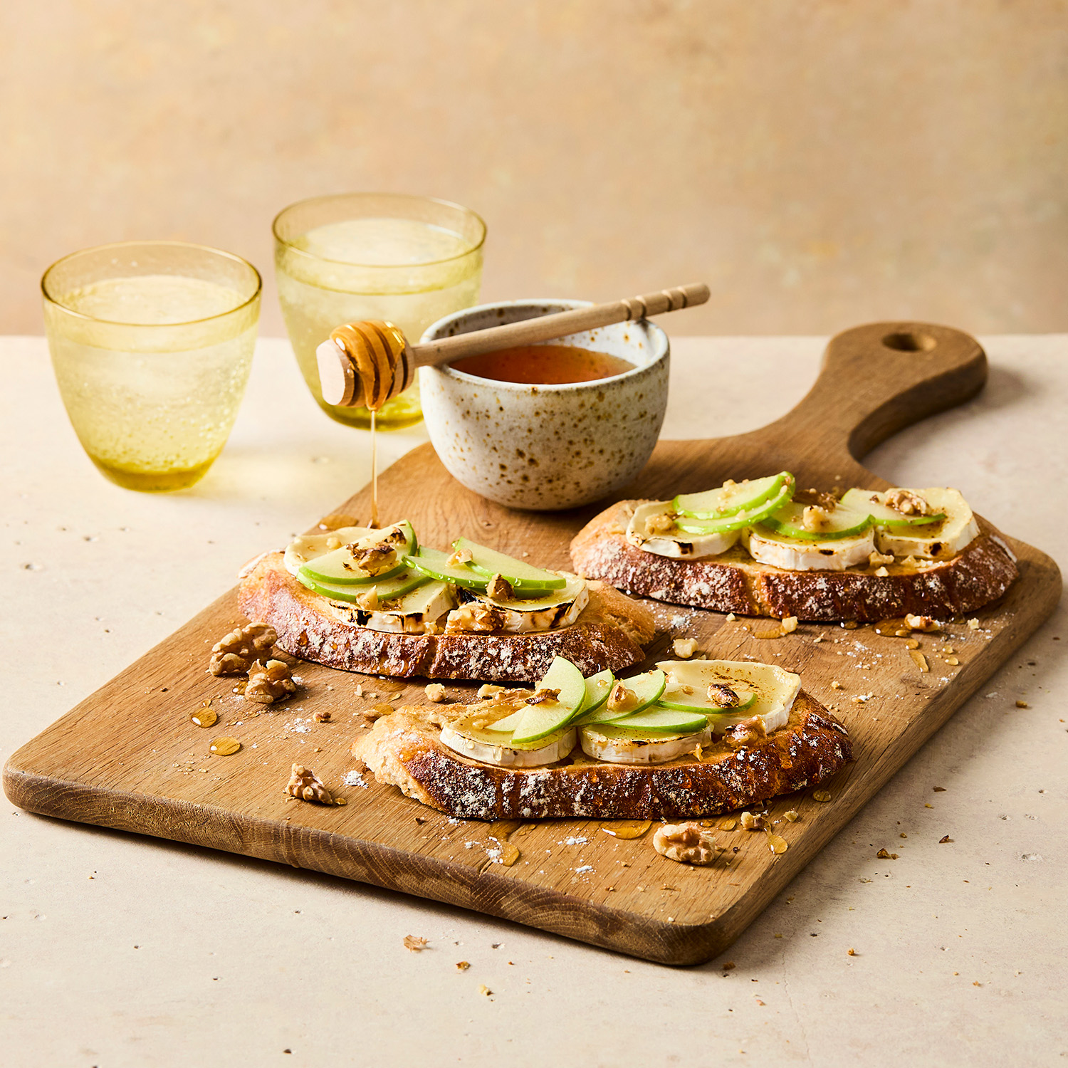Three slices of seeded sourdough on a wooden chopping board, topped with goats cheese, apple and walnut pieces. there is a pot of honey dripping onto the chopping board behind the toasts. In the background there are two glasses of water.