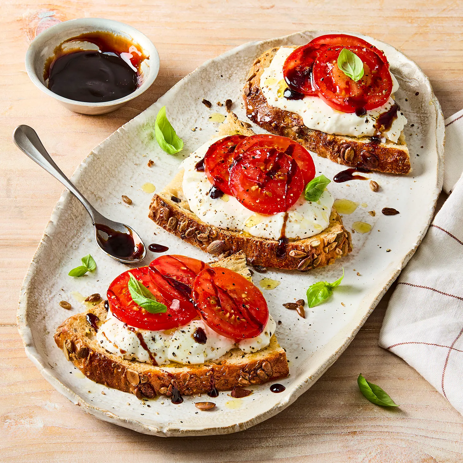 3 half slices of seeded sourdough on a plate topped with burrata cheese and tomato, drizzled with balsamic glaze and sprinkled with basil leaves. There is a spoon on the plate next to a bowl of extra balsamic glaze