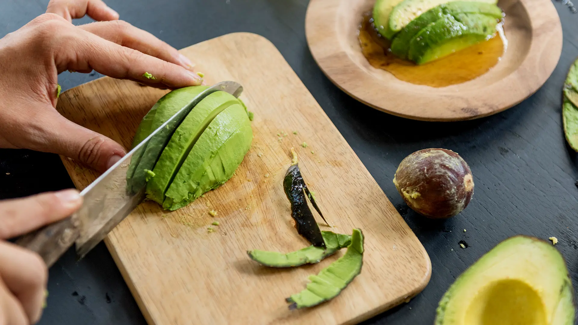 A top-down view of someone slicing half of an avocado on top of a wooden chopping board.