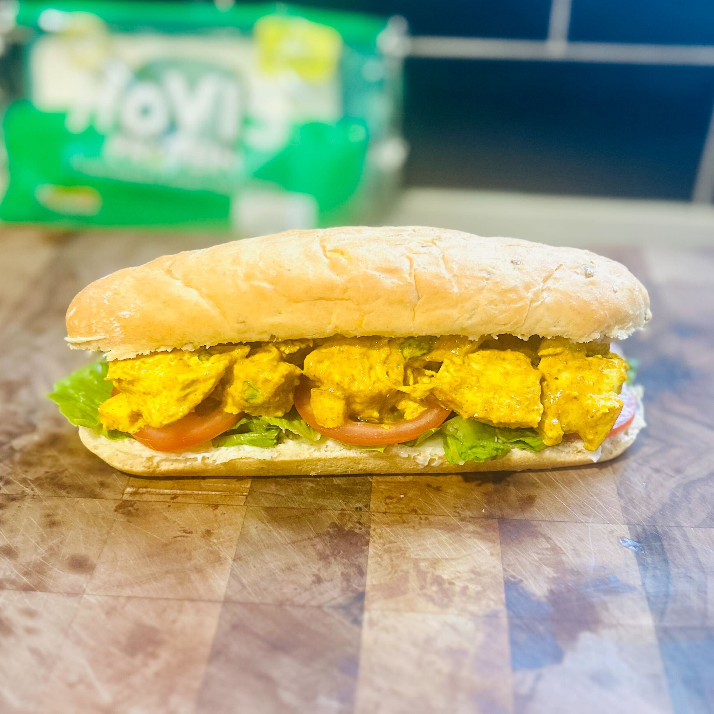 A side-view of a large, open-faced sub roll filled with Coronation Chicken. The sandwich is built on a wooden chopping board and contains shredded lettuce, sliced tomatoes, and a layer of creamy, yellow-curry-based Coronation Chicken. In the blurred background, a green bag of Hovis sub rolls is visible on a dark countertop with a tiled backsplash.
