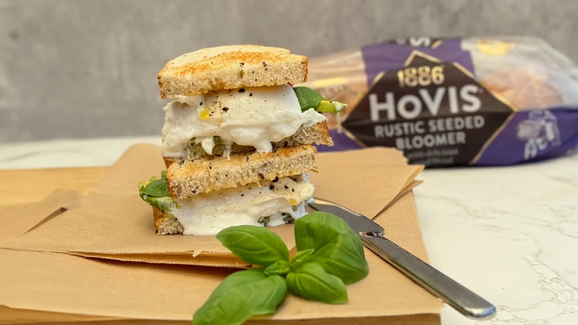 Two halves of a toasted Hovis Seeded Bloomer sandwich with creamy white cheese, basil leaves, avocado and small pieces of ground black pepper oozing out. In the foreground, additional basil leaves and a knife are visible.