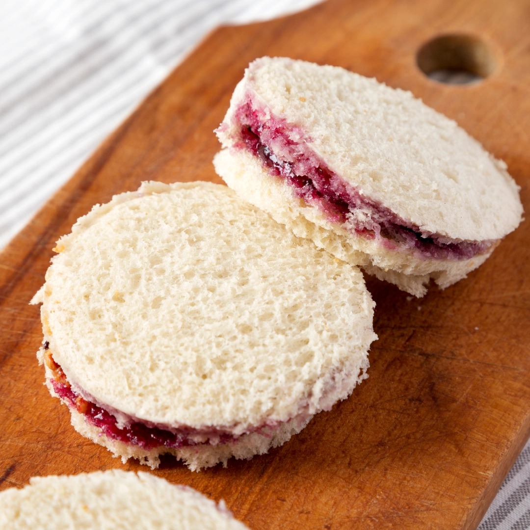 Two small, circular jam sandwiches made with Hovis Soft White Medium Sliced placed on a wooden chopping board, with the jam slightly visible between the slices of bread.