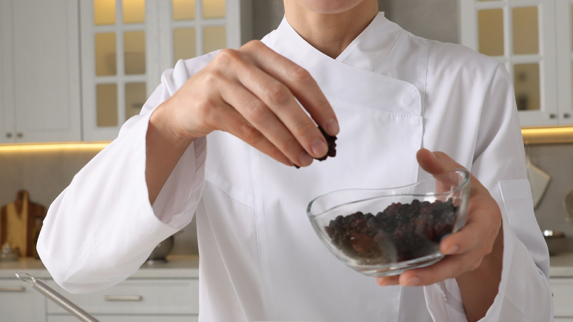Someone dressed as a chef, holding a small glass bowl full of raisins with one hand, while using the other to sprinkle the over a pot.
