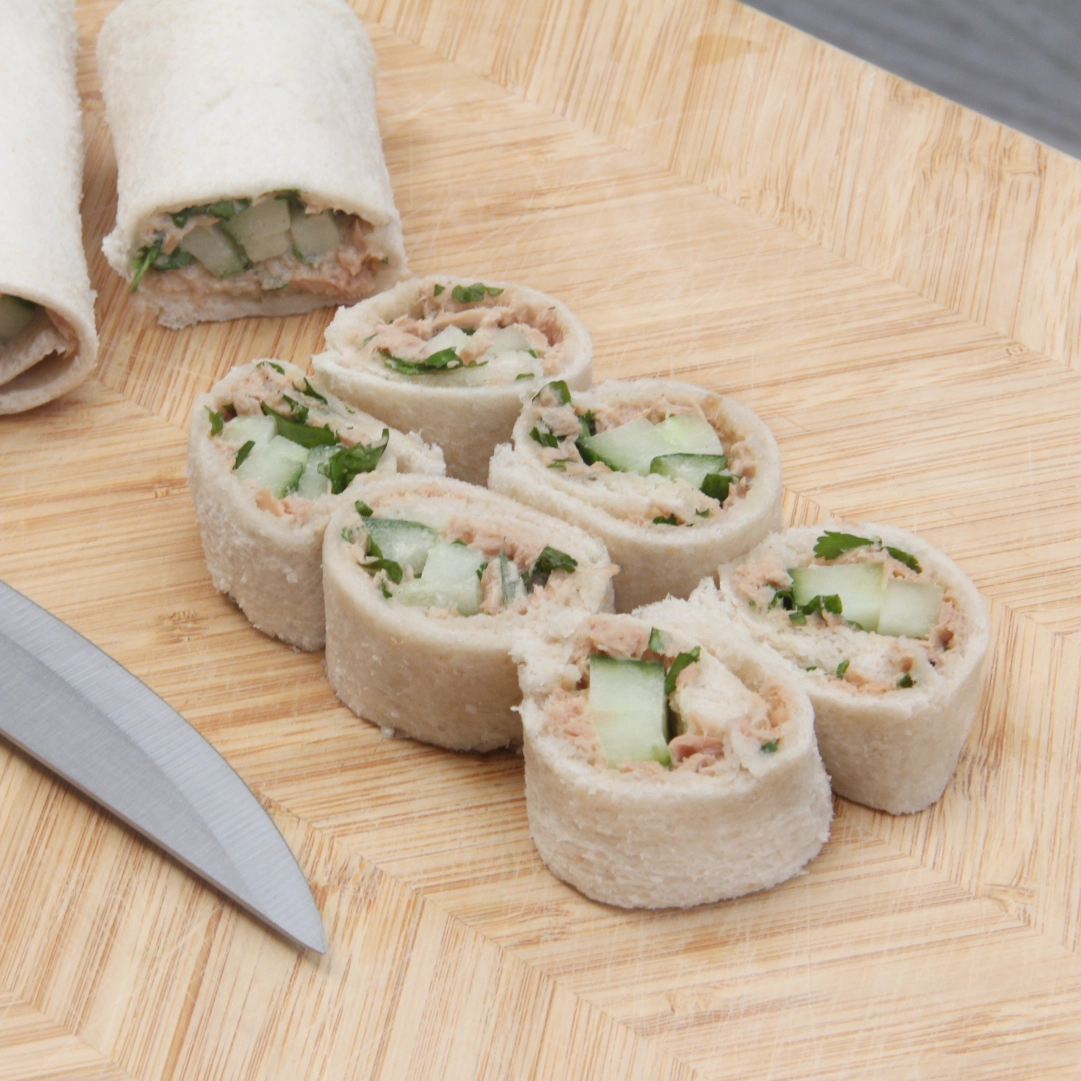 Hovis Soft White Thick Sliced wrapped around sliced cucumber and tuna mayo, cut into six bite-sized pieces resembling sushi, with two uncut Sandwich Sushi Roll in the background and a sharp knife.