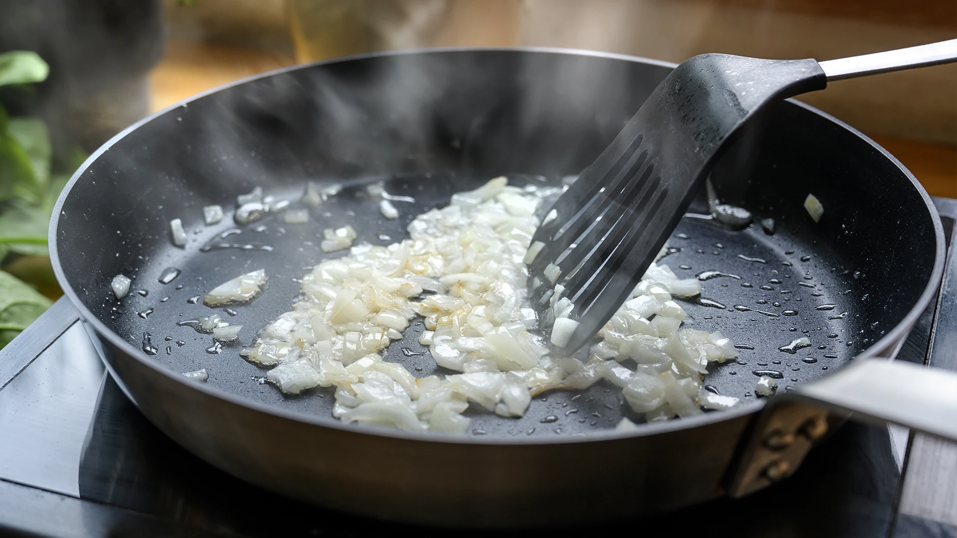 Sliced onions frying in a frying pan on a hob, being stirred with a black plastic spatula.