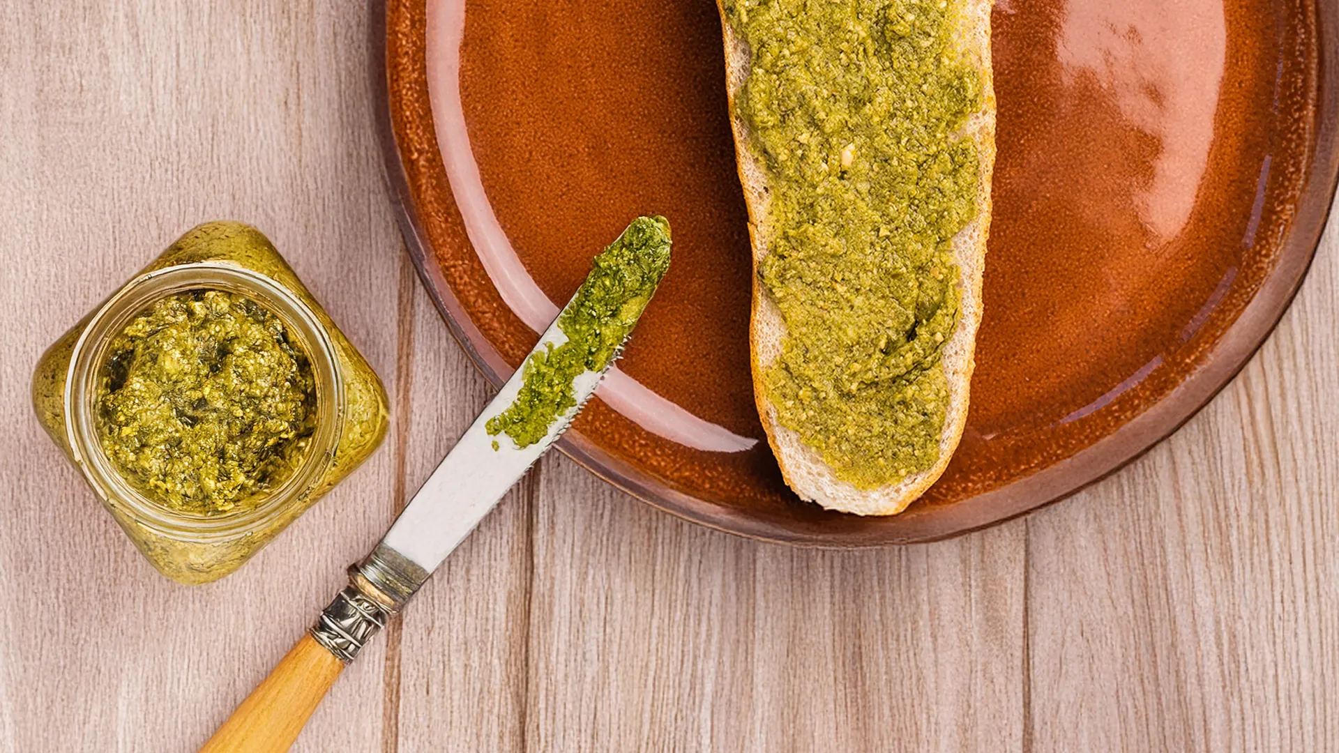 the bottom half of a sub roll sits on a red plate on a wooden kitchen counter with pesto spread on it, next to a knife covered in pesto and a jar of pesto
