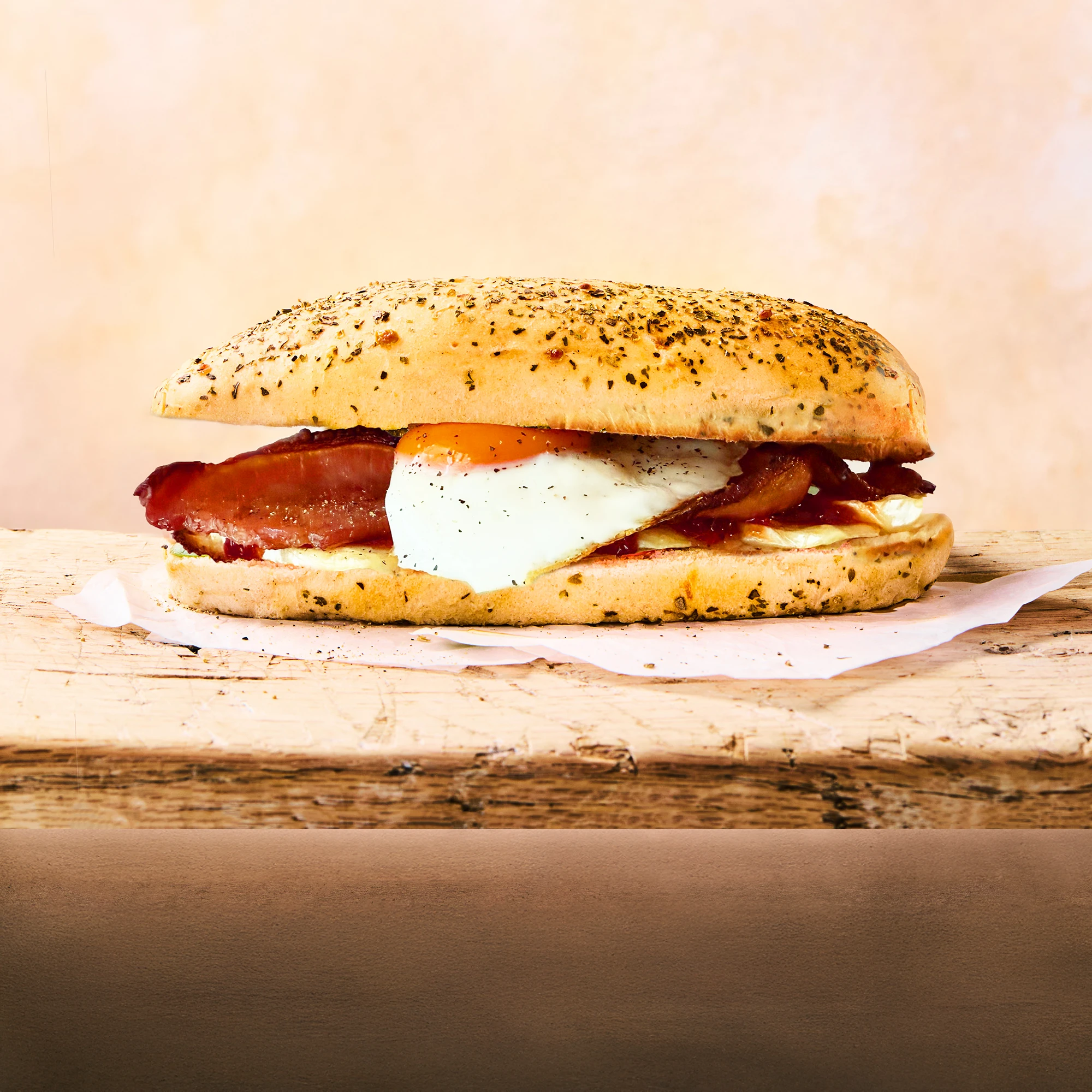 A complete melted brie and chilli jam sandwich sitting on parchment paper on a wooden surface with bacon, friend egg and chilli jam visible inside